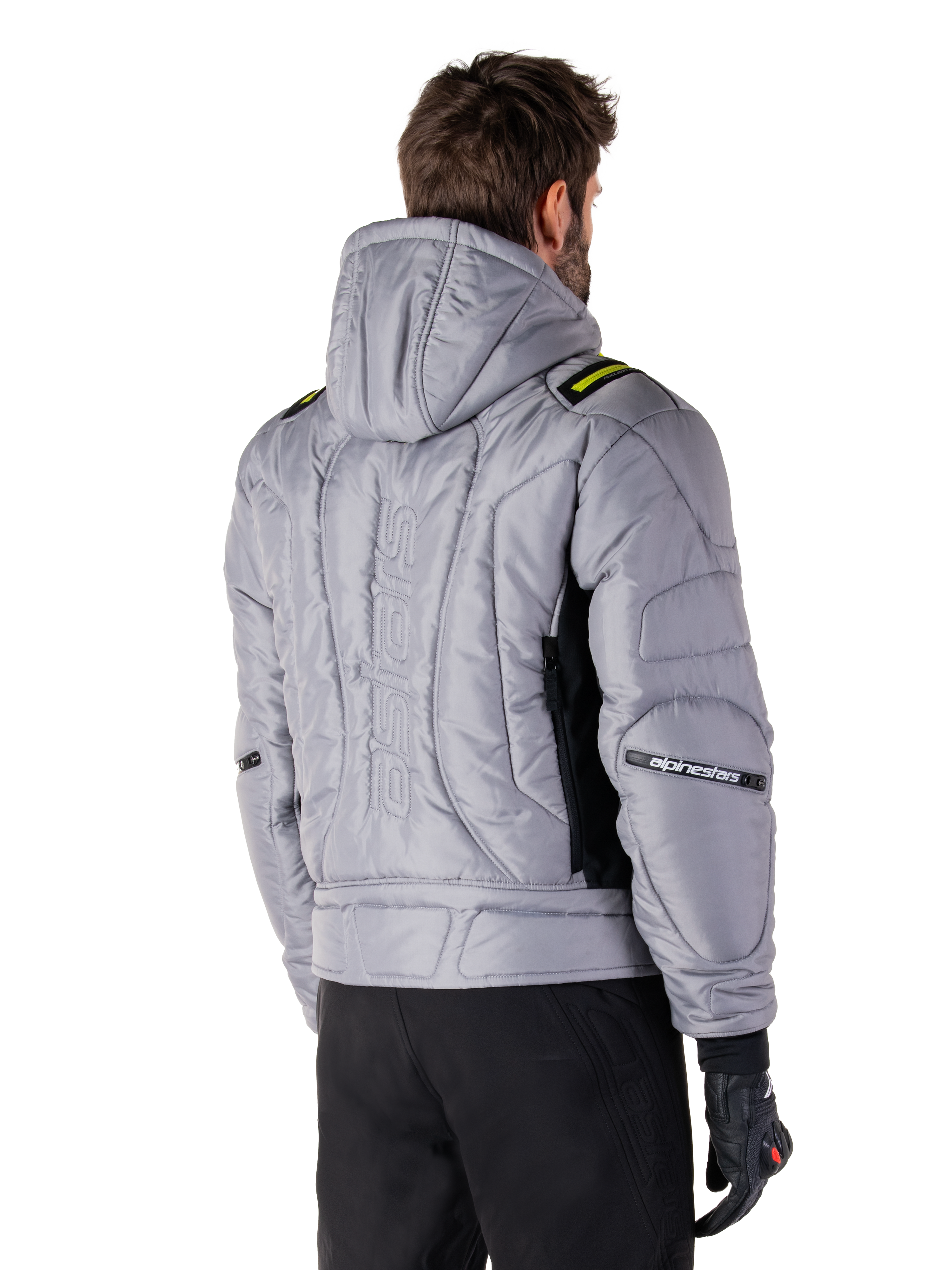 Alpinestars Mohobbs Waterproof Jas in Lunar Gray en Lime Punch, achteraanzicht op een model, gewatteerd bomberjack met verticaal astars-logo in reliëf op de rug, limegroene schouderaccenten, met capuchon, gedragen met zwarte motorbroek en handschoenen.