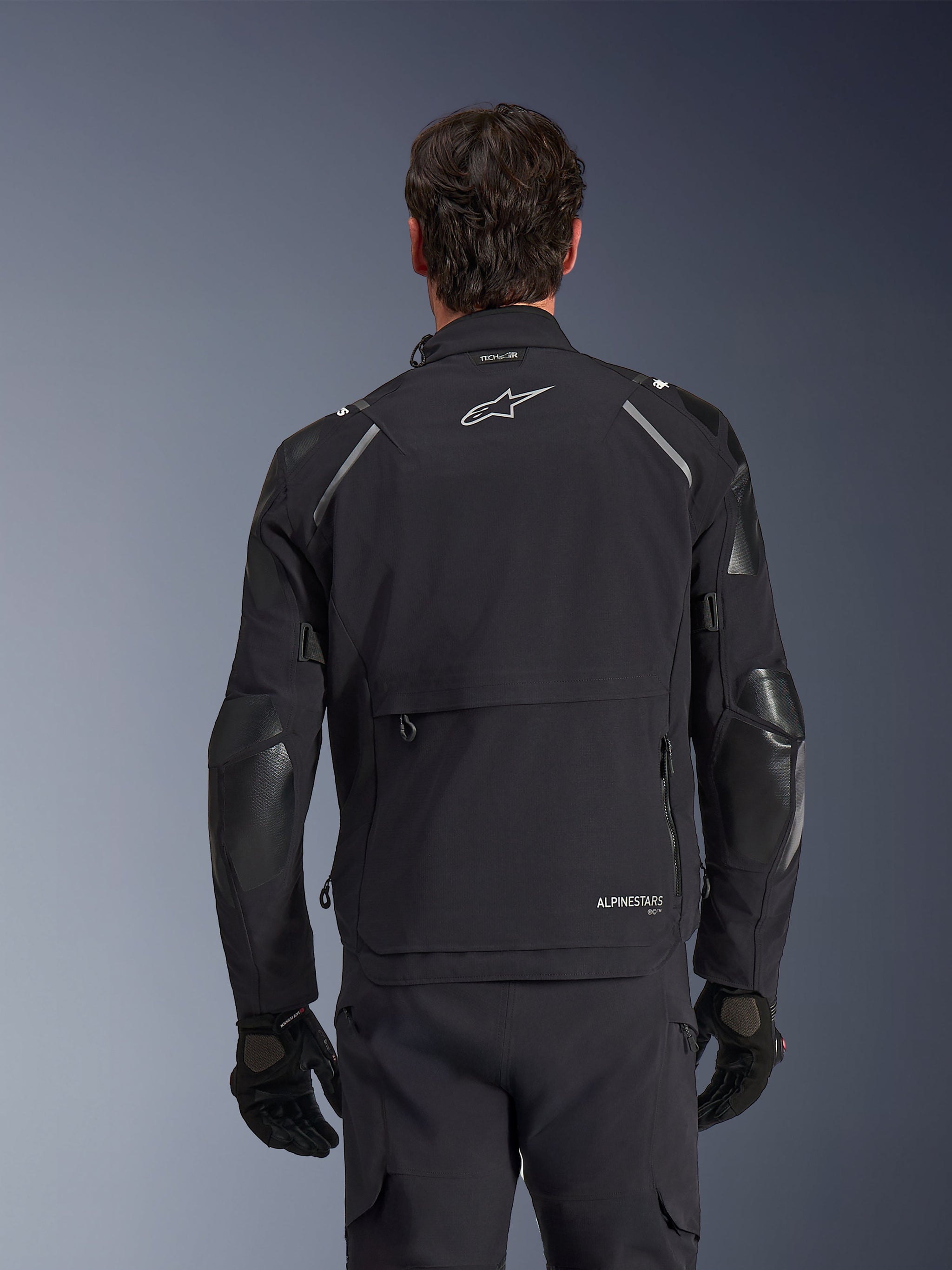 Alpinestars Andes Pro Drystar® XF jas, zwart en donkergrijs, achteraanzicht gedragen door een mannelijk model, met een groot opbergcompartiment aan de achterzijde, reflecterende schouderaccenten en merklogo's, gecombineerd met zwarte handschoenen en broek voor adventure touring