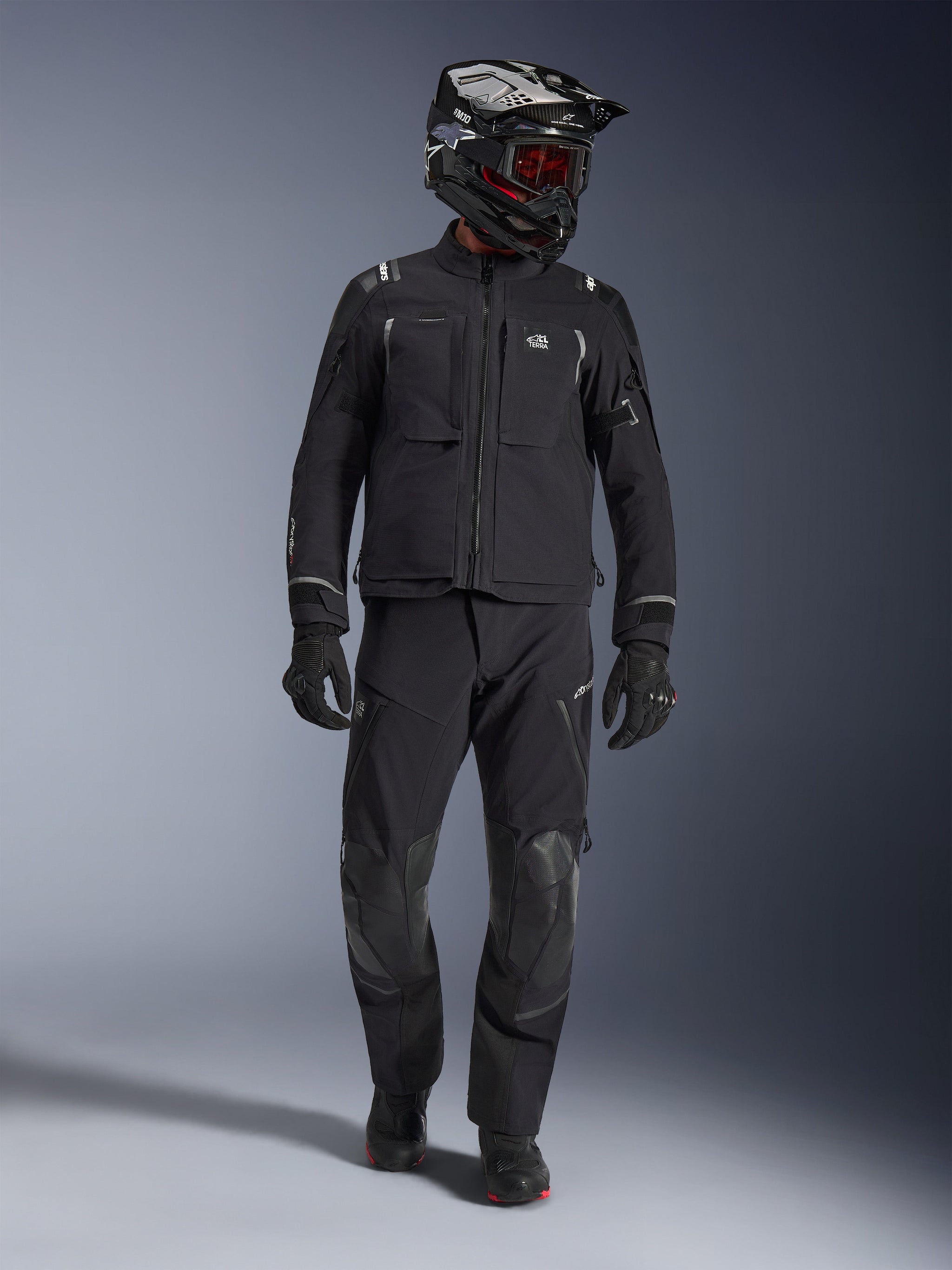 Adventure-rijder draagt de Alpinestars Andes Pro Drystar® XF Jas in zwart en donkergrijs, bijpassende zwarte adventure-broek, zwart-witte helm met rode bril, zwarte handschoenen en laarzen, voorzien van grote functionele zakken aan de voorzijde, reflecterende accenten en een technische 3-laags gelamineerde constructie voor touring in alle weersomstandigheden.