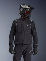 Adventure-rijder in de Alpinestars Andes Pro Drystar® XF Jas in zwart en donkergrijs, voorzien van All Terra-branding op de borst, gecombineerd met een bijpassende technische broek en een zwarte adventure-helm met bril, professionele touring-kledingset.