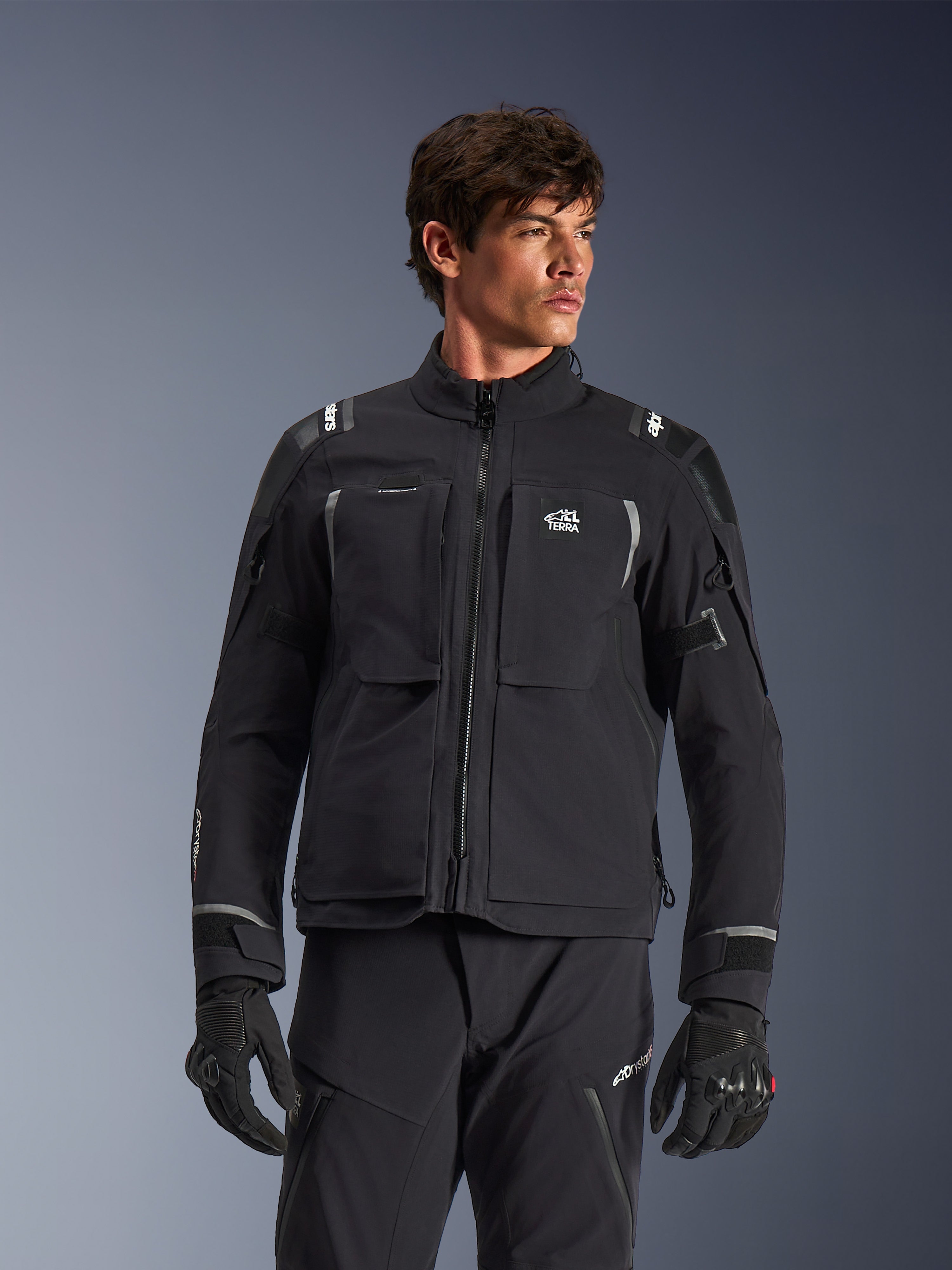 Alpinestars Andes Pro Drystar® XF Jacket, zwarte en donkergrijze adventure touring motorjas, gedragen met bijpassende broek en handschoenen, voorzien van een technische gelamineerde shell-constructie, ALL TERRA-logo op de borst, reflecterende details en verstelbare manchetten
