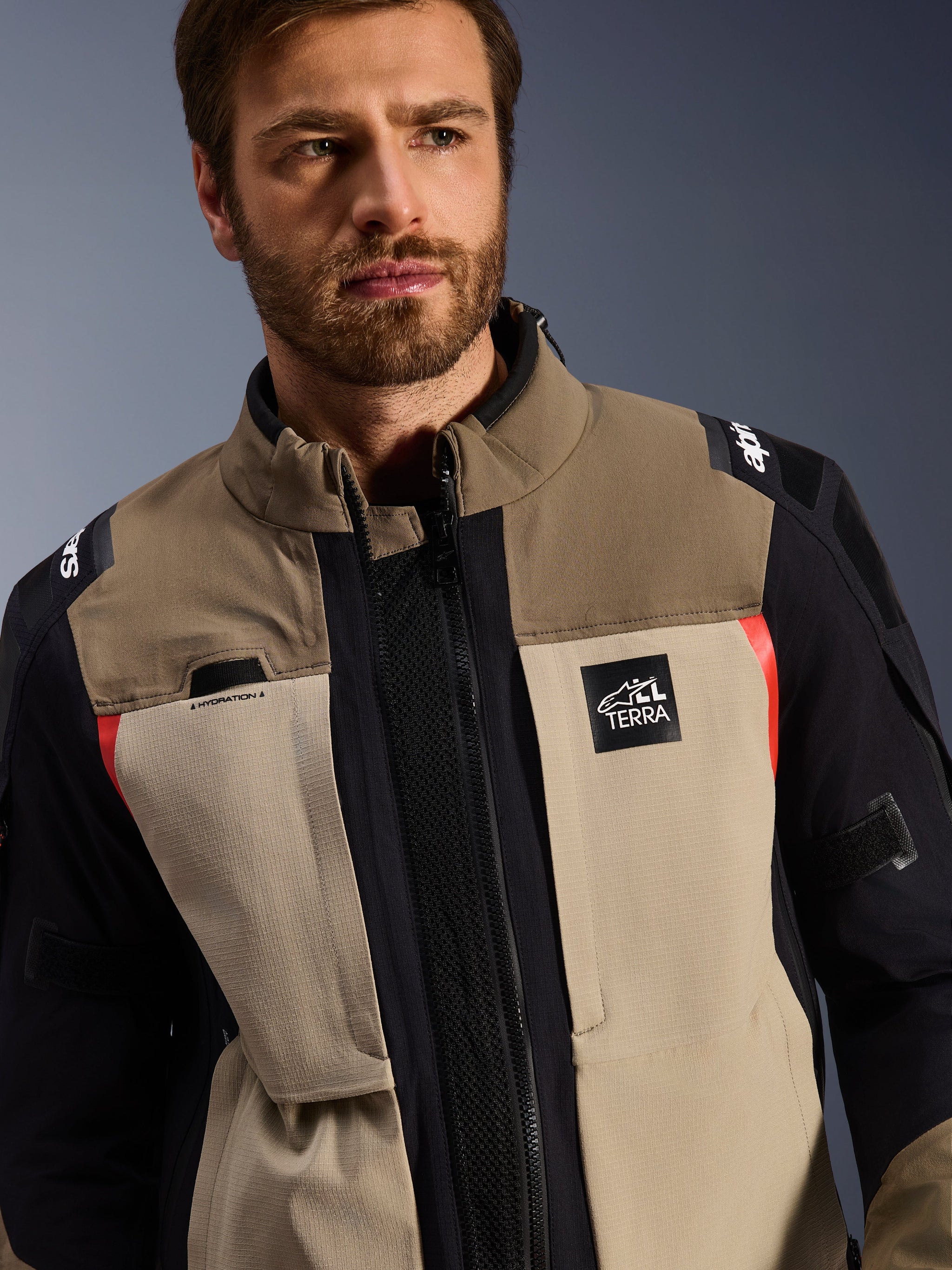 Alpinestars Andes Pro Drystar® XF Jacket in Walnut Aluminum, worn by a male model, adventure motorcycle jacket featuring a layered textile design in brown, light gray, and black with red accents, Alpinestars 'TERRA' logo on the chest, and visible technical details.