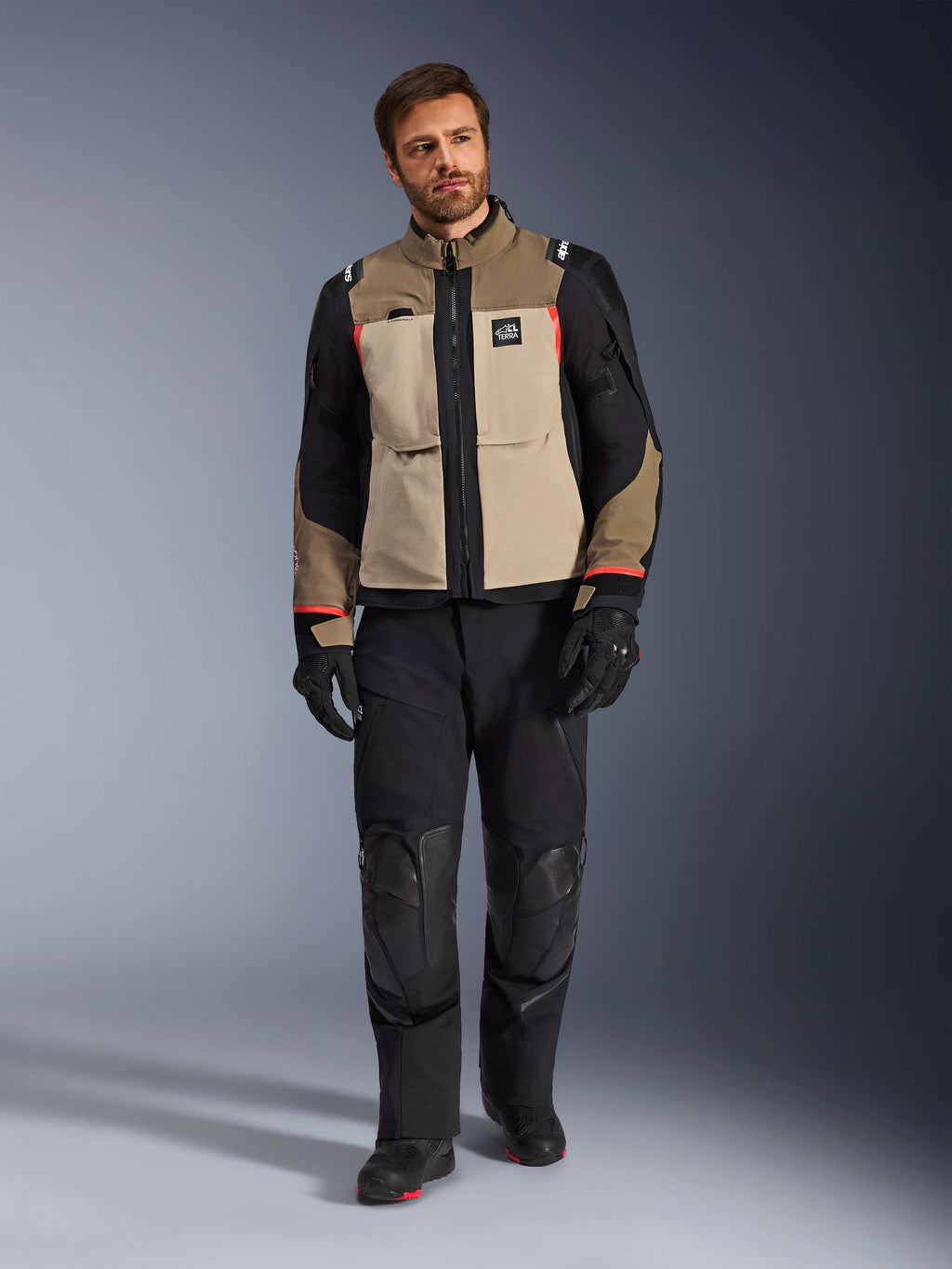 Mannelijk model draagt Alpinestars Andes Pro Drystar® XF Jacket in Walnut Aluminum, zandkleurige en zwarte adventure touring jas met rode accenten, zwarte motorbroek, handschoenen en laarzen, studio-opname van het volledige lichaam, beschermende motorkleding