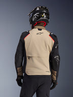 Alpinestars Andes Pro Drystar® XF Jas in Walnut Aluminum, achteraanzicht, beige achterpaneel met grote cargozak, zwarte mouwen met versterkte panelen, Alpinestars-logo op de bovenrug, gecombineerd met een zwarte helm en handschoenen voor adventure touring.