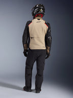Rijder van achteren in de Alpinestars Andes Pro Drystar® XF Jas in Walnut Aluminum, beige rugpand met zwarte mouwen en rode accenten, gecombineerd met een zwarte technische adventure-broek en een motorhelm, hoogwaardige all-weather touringuitrusting.