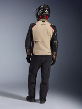Rider from the back wearing the Alpinestars Andes Pro Drystar® XF Jacket in Walnut Aluminum, tan back panel with black sleeves and red accents, paired with black technical adventure pants and a motorcycle helmet, premium all-weather touring gear.