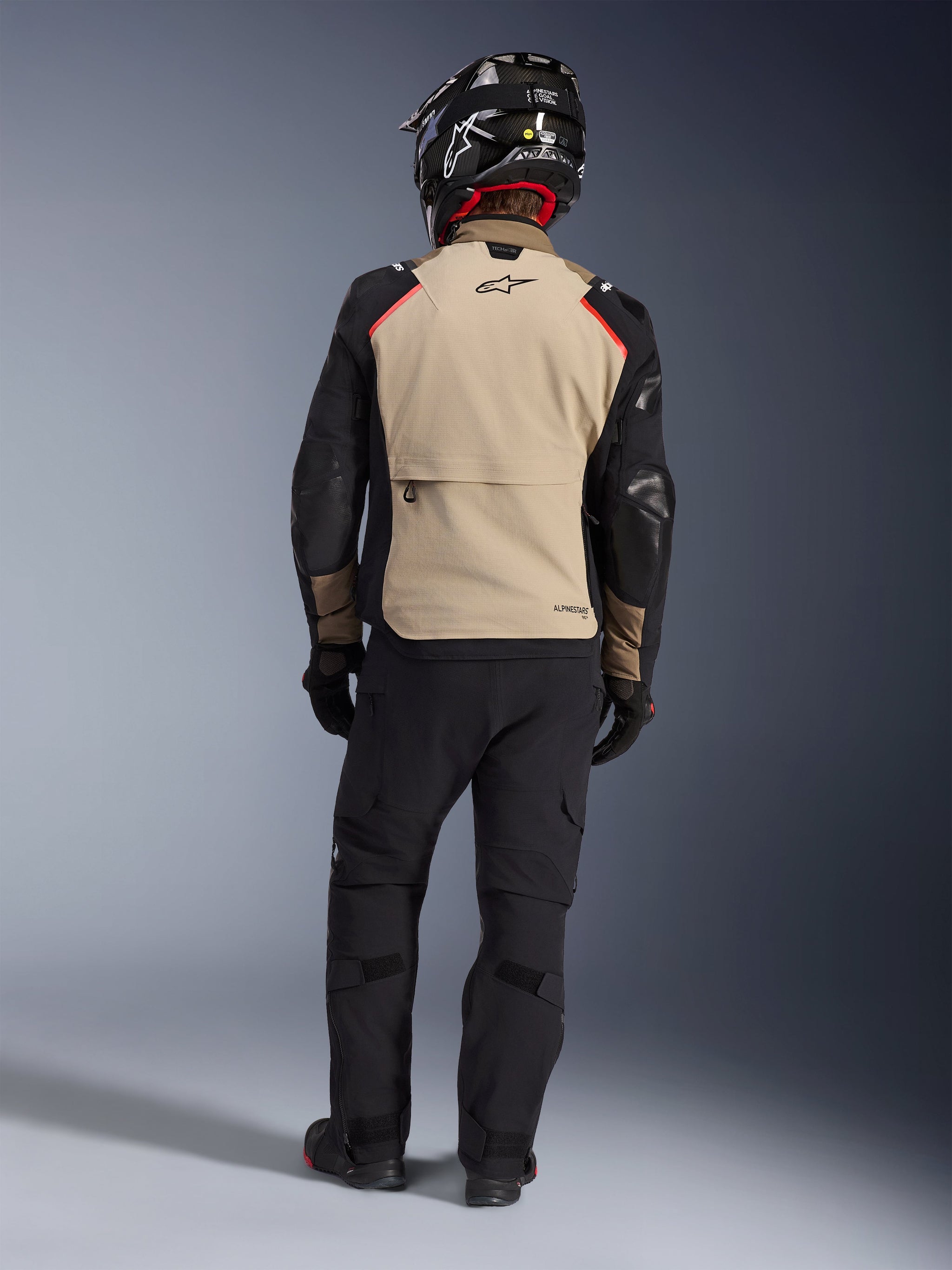 Rider from the back wearing the Alpinestars Andes Pro Drystar® XF Jacket in Walnut Aluminum, tan back panel with black sleeves and red accents, paired with black technical adventure pants and a motorcycle helmet, premium all-weather touring gear.