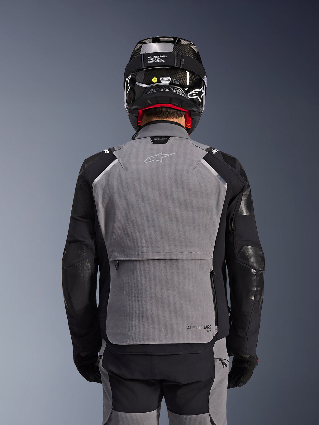 Alpinestars Andes Pro Drystar® XF Jas, donkergrijs en zwart, achteraanzicht met grote opbergzak op de rug en Tech-Air branding, gedragen door een motorrijder met een zwarte helm en bijpassende motorbroek, professionele adventure touring uitrusting