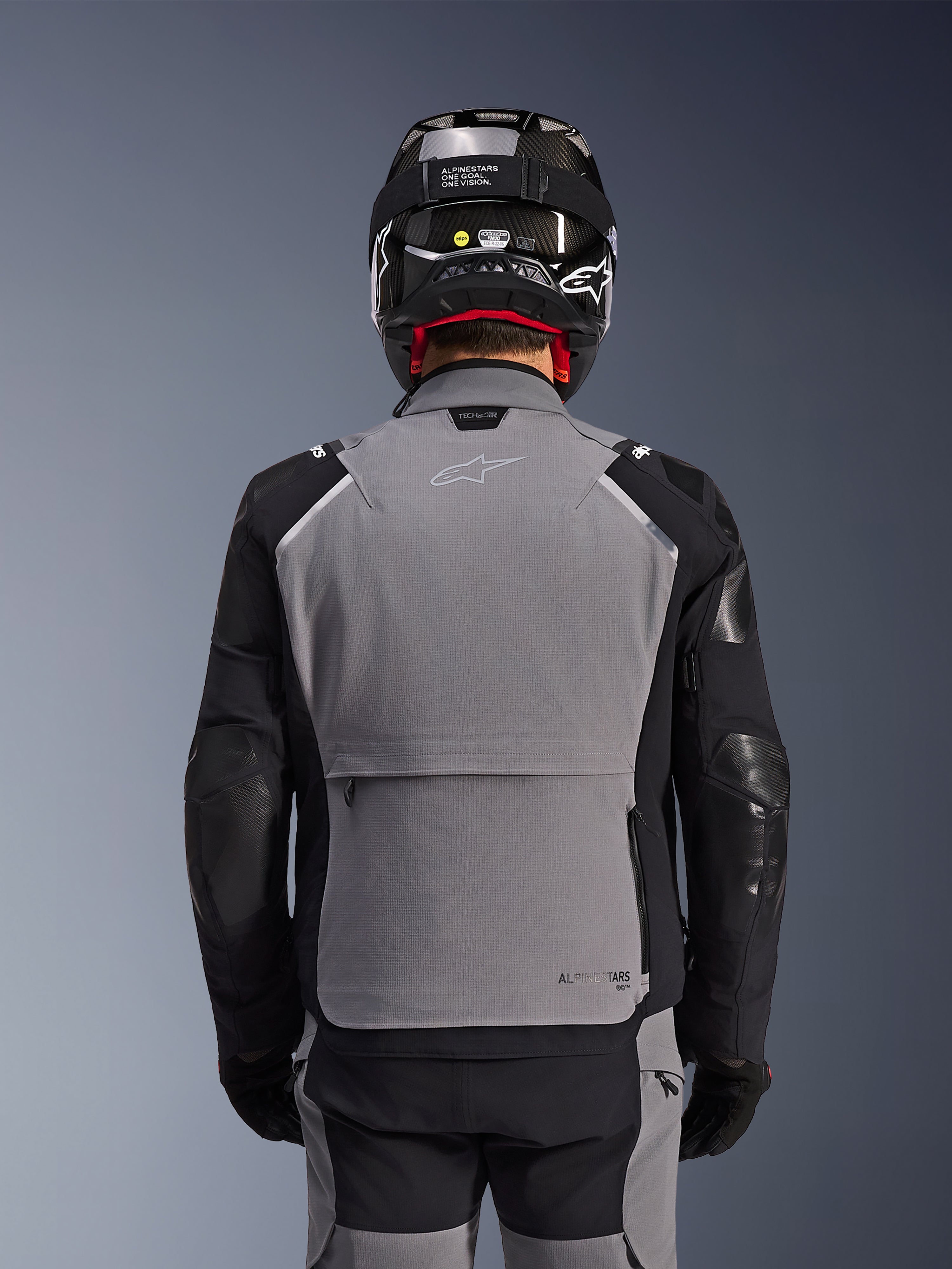 Alpinestars Andes Pro Drystar® XF Jas, donkergrijs en zwart, achteraanzicht met grote opbergzak op de rug en Tech-Air branding, gedragen door een motorrijder met een zwarte helm en bijpassende motorbroek, professionele adventure touring uitrusting