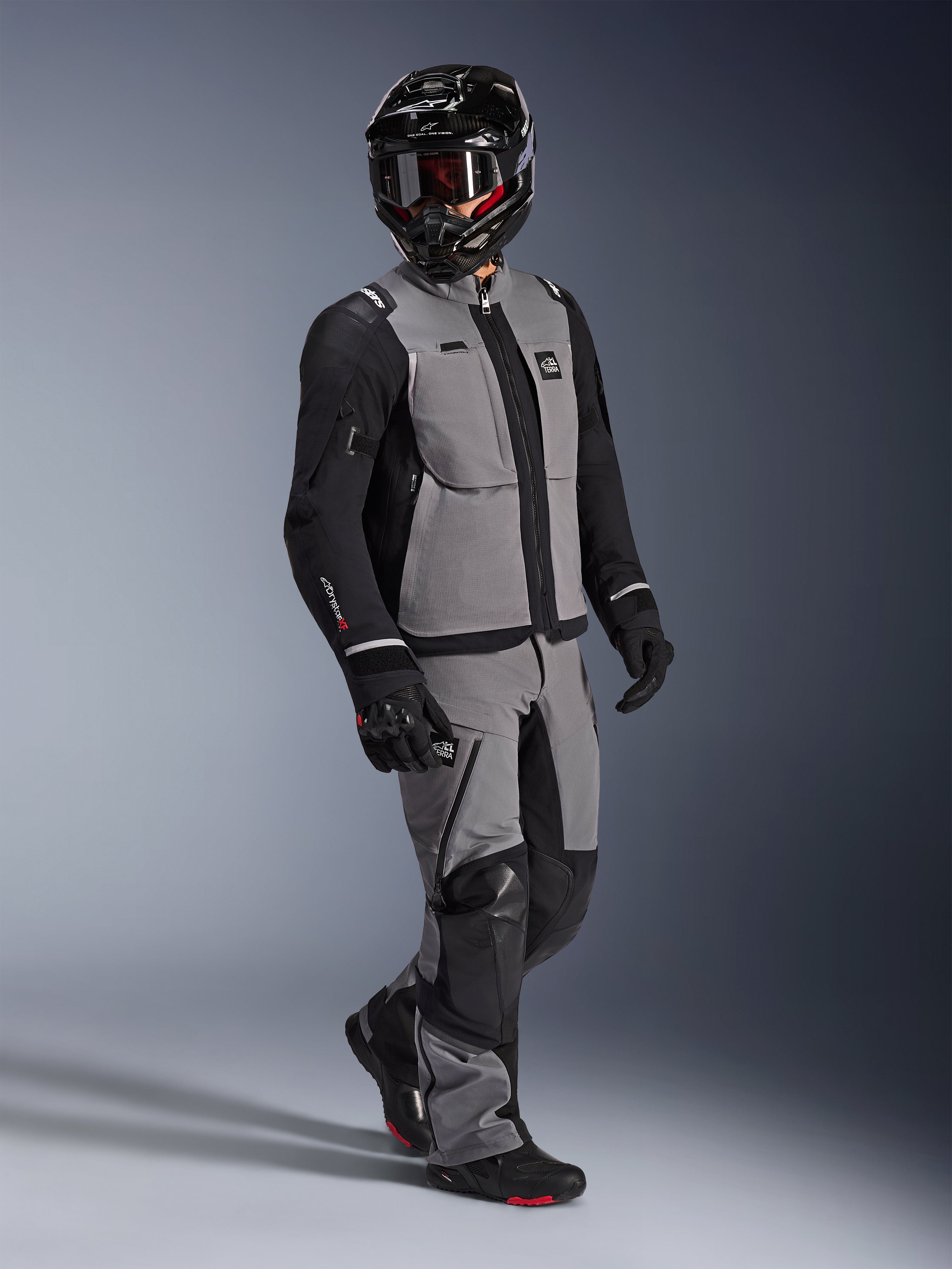 Adventure-rijder met de Alpinestars Andes Pro Drystar® XF Jas in donkergrijs en zwart, bijpassende grijze en zwarte adventure-broek, zwarte integraalhelm met motorbril en zwarte motorhandschoenen, studio-opname voor professionele touringuitrusting.