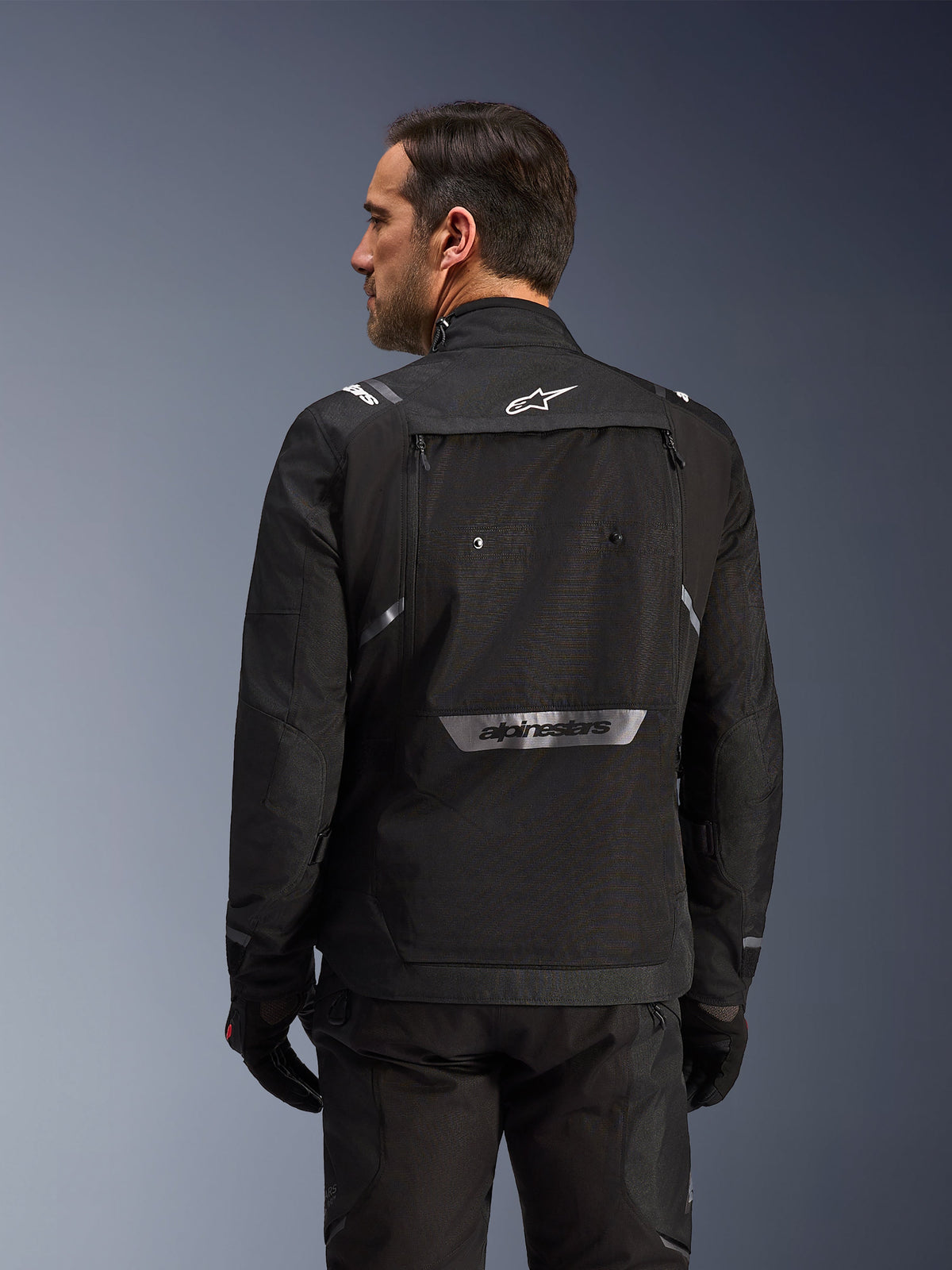 Motorrijder van achteren gezien die de Alpinestars Cusco Drystar® Jas in het zwart draagt, textiele adventure touring jas met grote opbergzak op de rug en reflecterende merkdetails, gecombineerd met bijpassende zwarte broek en handschoenen