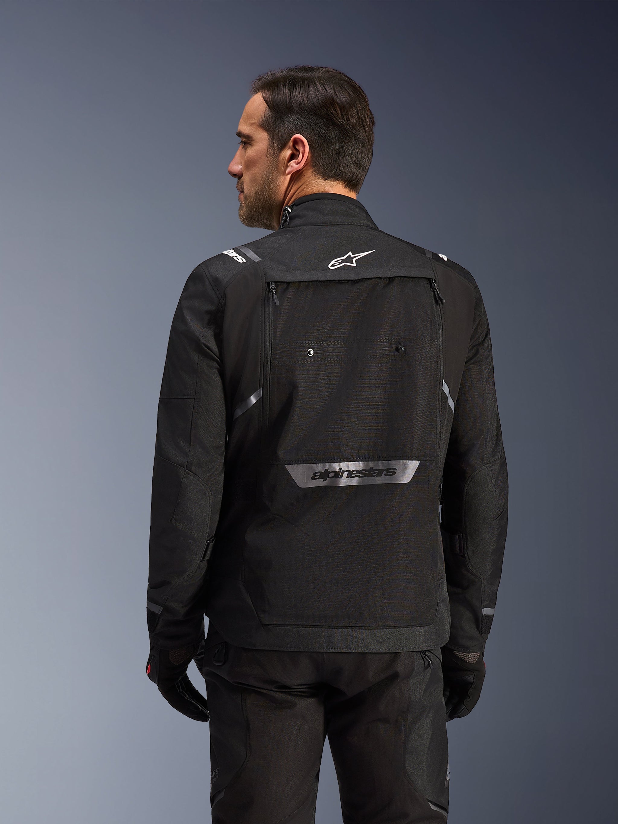 Rider from behind wearing Alpinestars Cusco Drystar® Jacket in Black, textile adventure touring jacket with large rear storage pocket and reflective branding, paired with matching black pants and gloves
