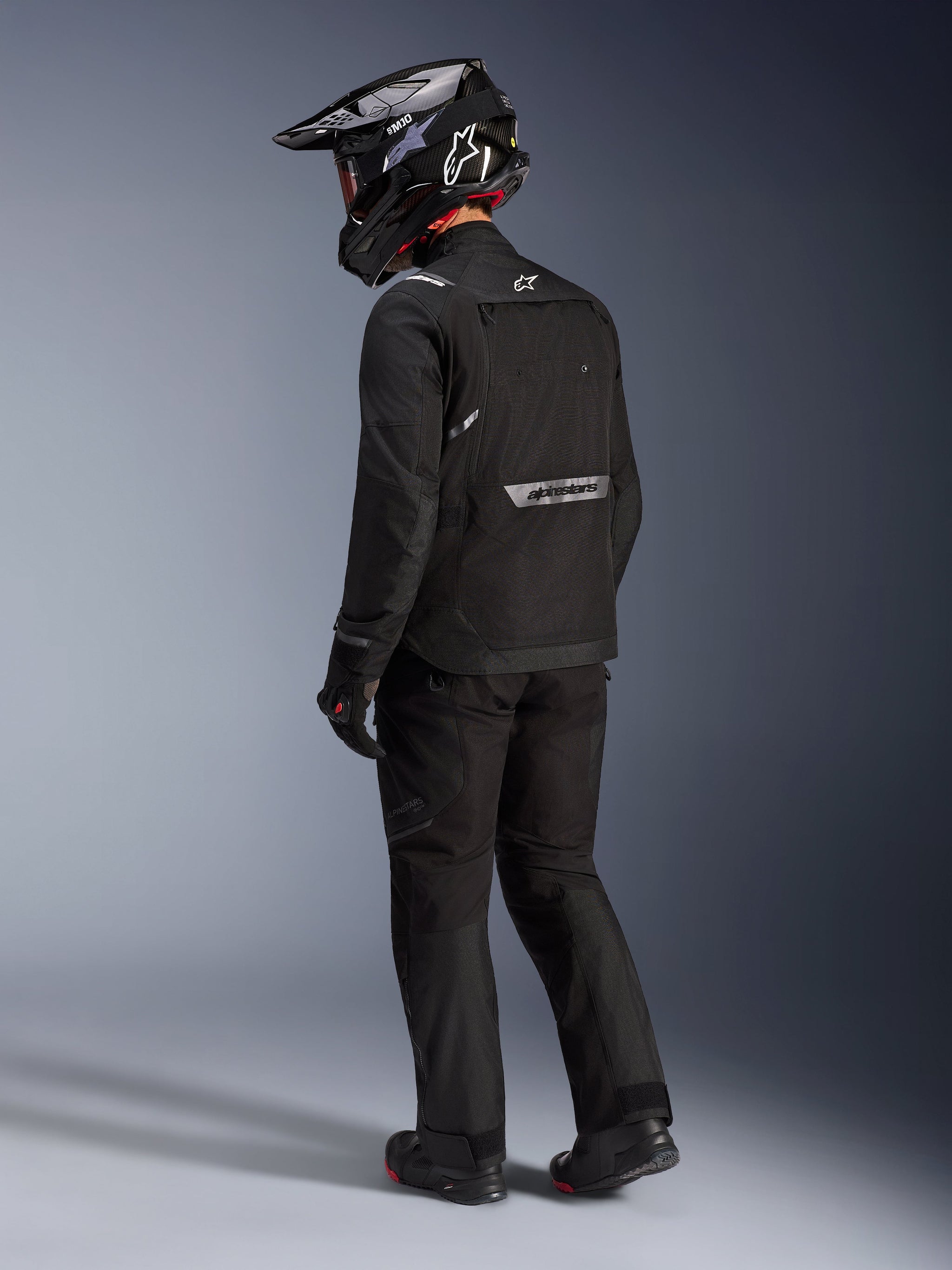 Alpinestars Cusco Drystar® Jacket in Black, rear-side view of a rider wearing a full adventure touring kit including matching black pants and gloves, black adventure helmet, textile construction with reflective branding on the back