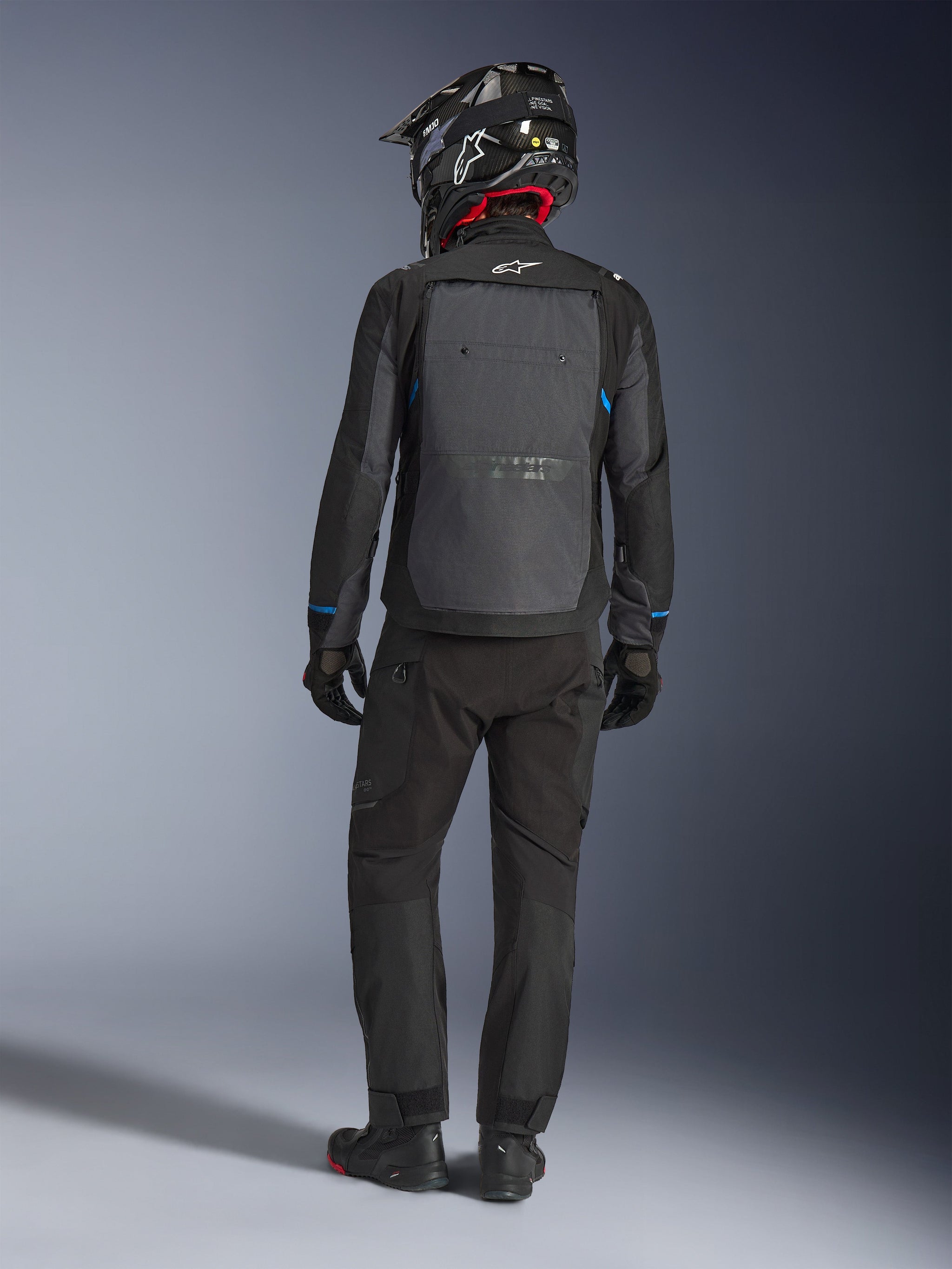 Alpinestars Cusco Drystar® Jacket, zwart en donkergrijs, achteraanzicht van een motorrijder die volledige adventure touring-uitrusting draagt, groot grijs rugpaneel met Alpinestars-logo, blauwe accenten op de mouwen, gedragen met bijpassende zwarte broek en helm.