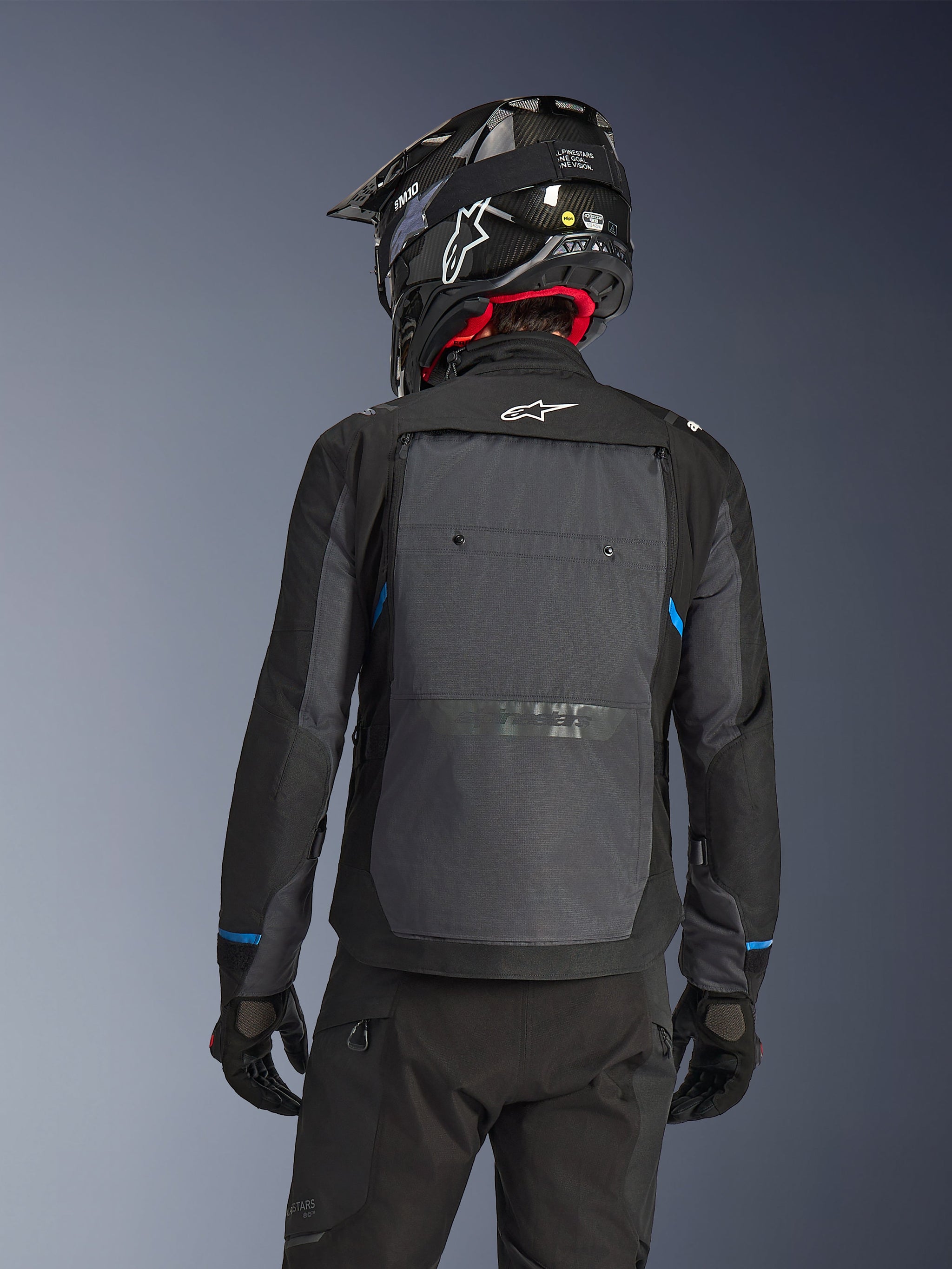 Motorrijder van achteren die de Alpinestars Cusco Drystar® Jas in Black Deep Gray draagt, met een groot grijs ventilatiepaneel op de rug en een wit logo, gecombineerd met een zwarte off-road helm en zwarte motorkleding voor adventure touring.