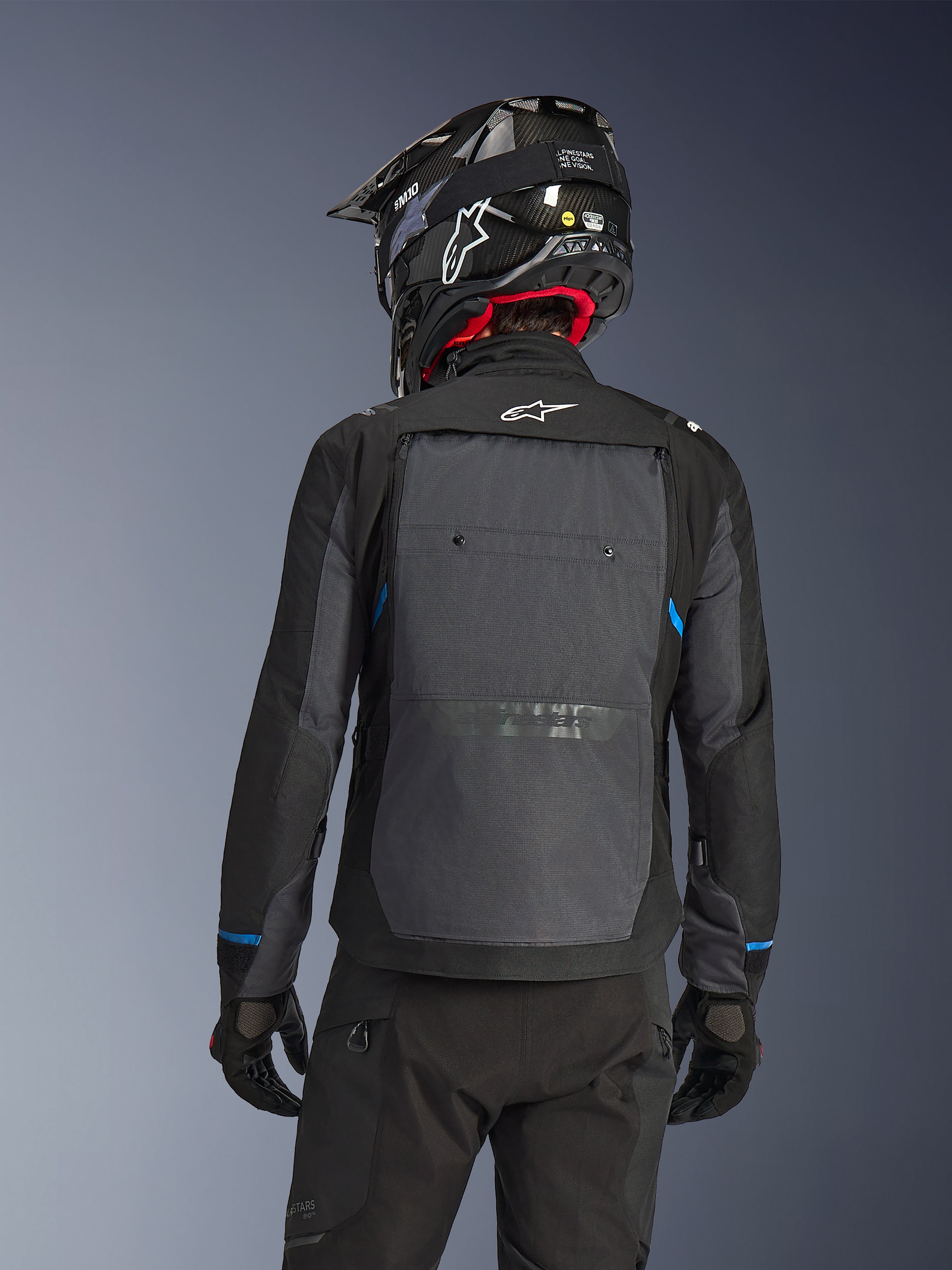 Rider from behind wearing the Alpinestars Cusco Drystar® Jacket in Black and Deep Gray, featuring a large gray back ventilation panel and white logo, paired with a black off-road helmet and black riding gear for adventure touring.