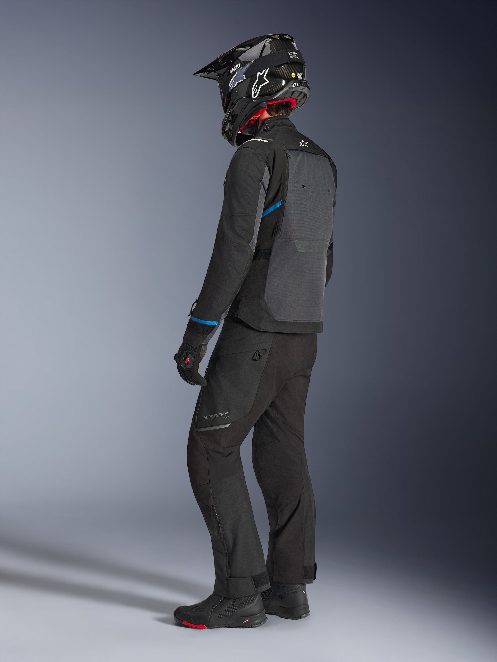Motorrijder die de Alpinestars Cusco Drystar® jas draagt in zwart en diepgrijs met blauwe accenten, gecombineerd met een zwarte motorbroek, een carbon SM10-helm en zwarte handschoenen, achteraanzicht met het grote ventilatiepaneel en adventure touring-ontwerp.