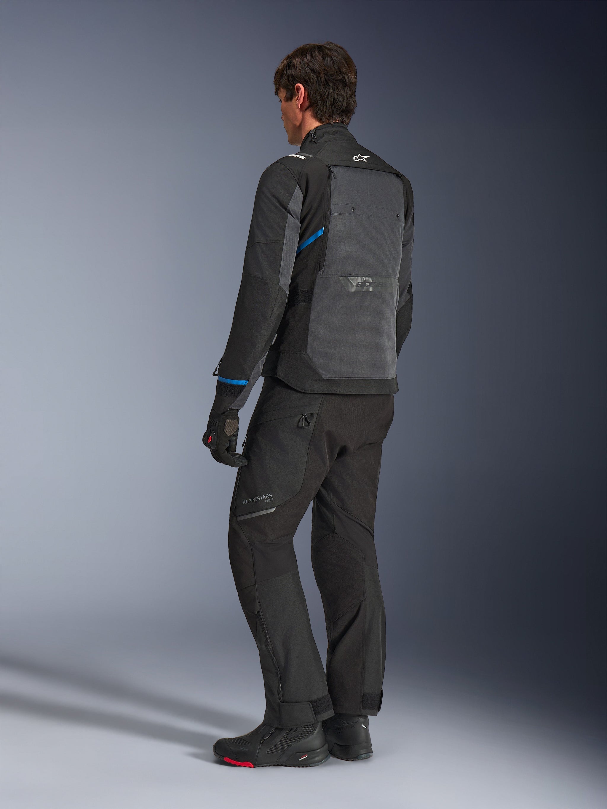Alpinestars Cusco Drystar® Jas, zwart en diepgrijs, driekwart achteraanzicht met groot ventilatiepaneel op de rug en blauwe accenten op de mouwen, gedragen met zwarte motorbroek en laarzen voor een complete touring outfit.