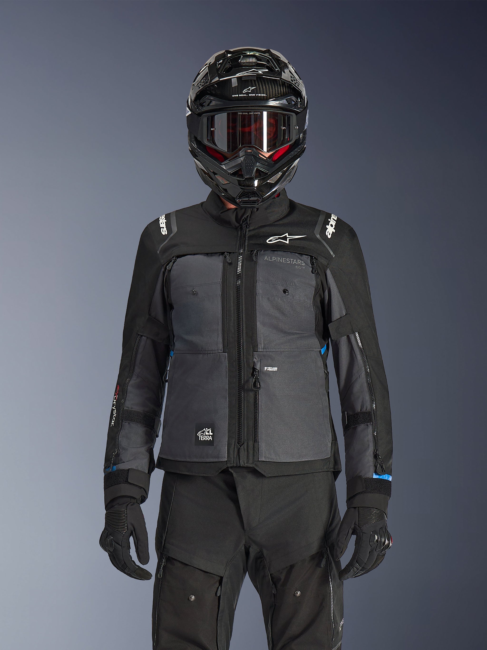 Alpinestars Cusco Drystar® Jas, zwart en donkergrijs, adventure touring motorjas gedragen met bijpassende zwarte technische broek en een dual-sport helm, tweekleurig ontwerp met ripstop-panelen en Alpinestars-branding