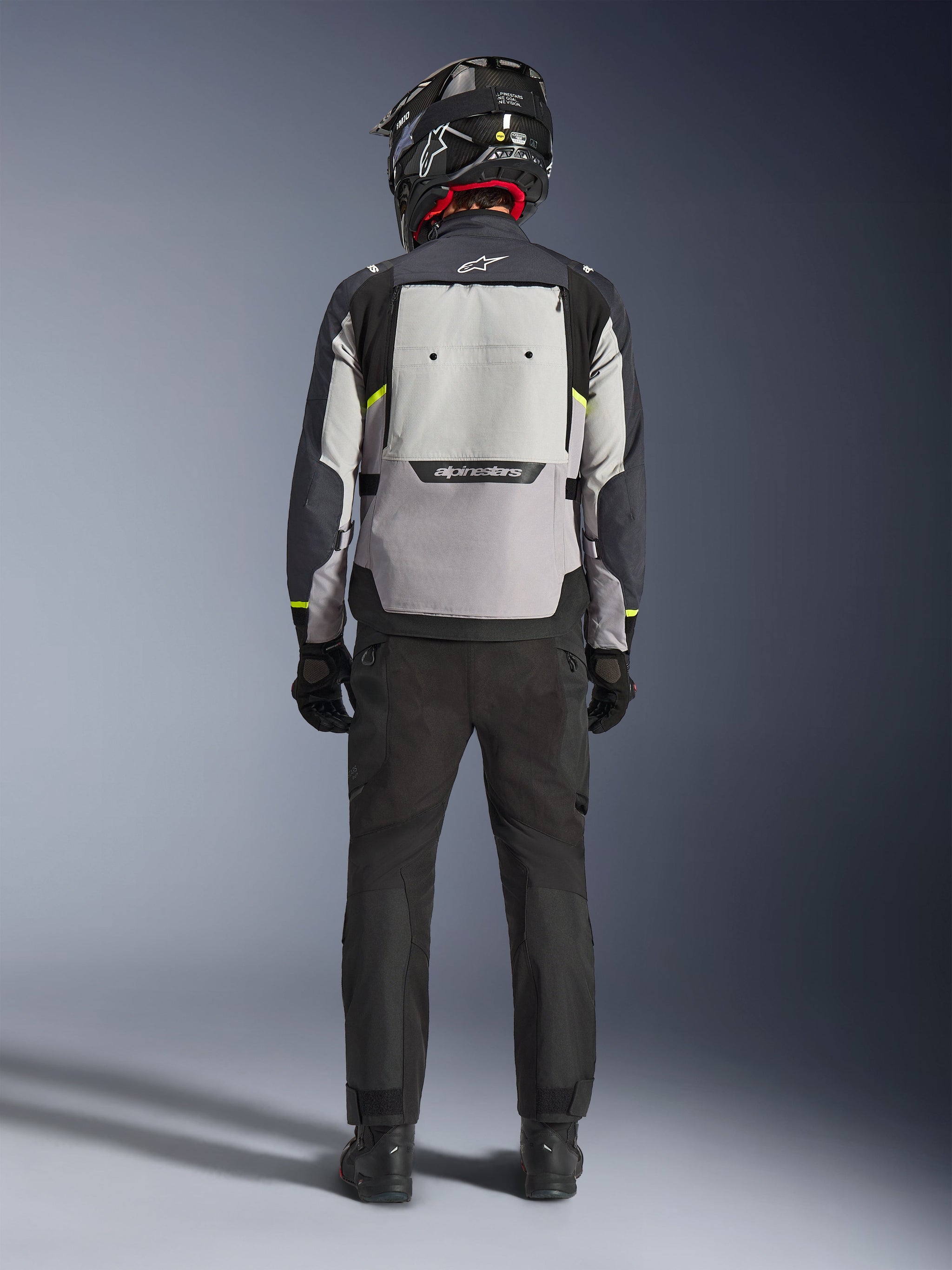 Motorrijder van achteren gezien met de Alpinestars Cusco Drystar® Jas in Ice Gray, Mid Gray en Deep Gray, voorzien van een groot ventilatiepaneel op de rug en Alpinestars-branding, gecombineerd met een zwarte motorbroek en een zwarte helm, adventure touring motorkleding