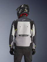 Alpinestars Cusco Drystar® Jacket, achteraanzicht, Ice Gray, Mid Gray en Deep Gray, voorzien van een groot ventilatiepaneel op de rug en felgele accenten, gecombineerd met een zwarte adventure-helm en zwarte broek.