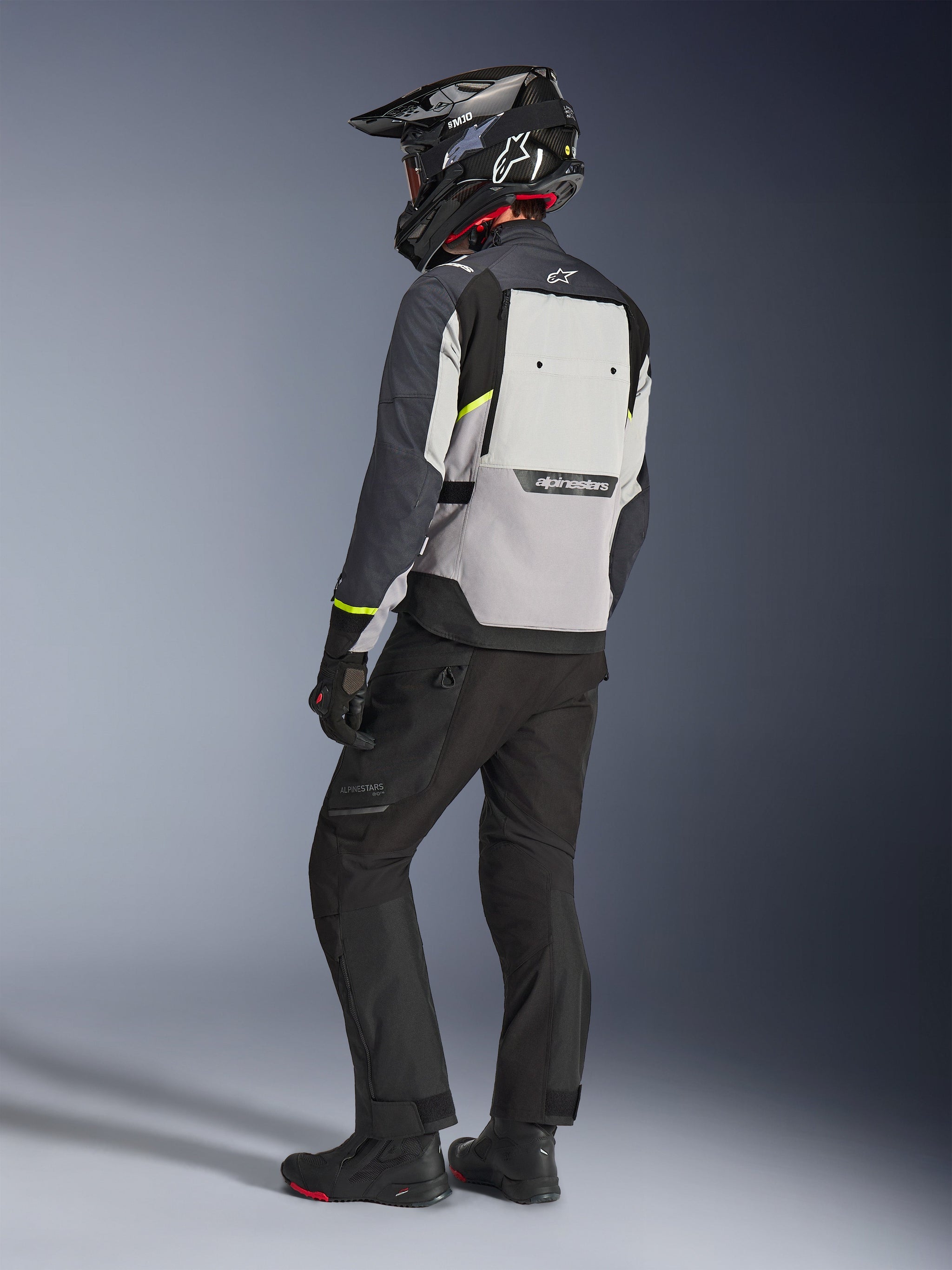 Alpinestars Cusco Drystar® Jas in Ice Gray, Mid Gray en Deep Gray, achteraanzicht op een motorrijder. Voorzien van fluorescerend gele accenten, een groot ventilatiepaneel op de rug met merkaanduiding en een duurzame textielconstructie. Gedragen met een zwarte motorbroek en een adventure-helm voor een complete touring-look.