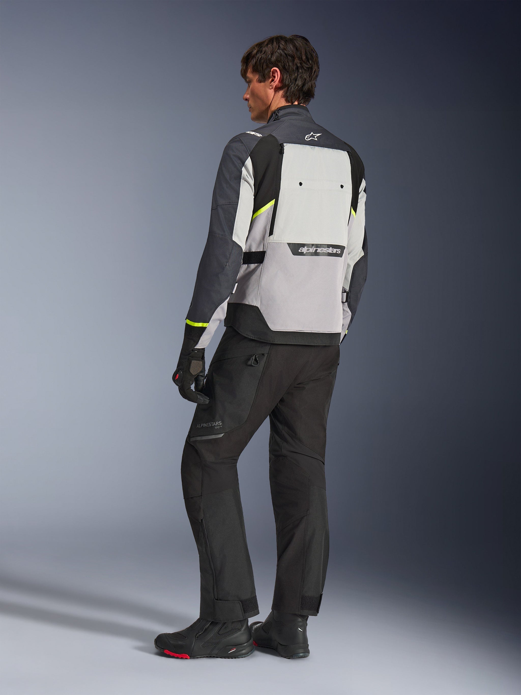 Alpinestars Cusco Drystar® Jas in Ice Gray, Mid Gray en Deep Gray, achteraanzicht op model, adventure touring jas met neongele accenten, gedragen met zwarte motorbroek en laarzen, voorzien van een groot ventilatiepaneel op de rug en Alpinestars-branding.