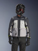 Alpinestars Cusco Drystar® Jas, adventure touring motorjas, Ice Gray, Mid Gray en Deep Gray, met grijze panelen in verschillende tinten, ventilatieopeningen met ritssluiting op de borst en Alpinestars branding, gedragen door een motorrijder met een zwarte helm en motorbril.