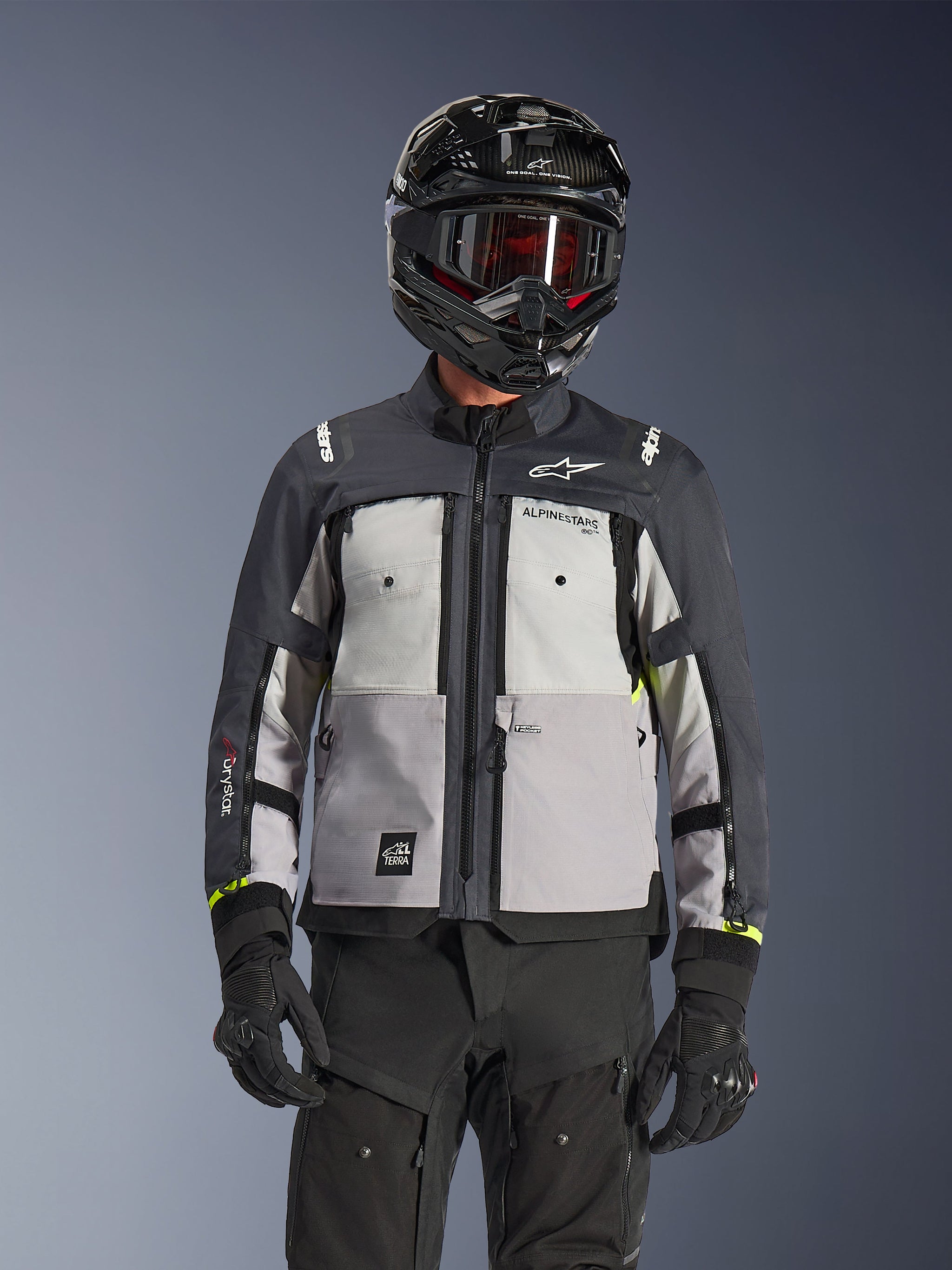 Alpinestars Cusco Drystar® Jas, adventure touring motorjas, Ice Gray, Mid Gray en Deep Gray, met grijze panelen in verschillende tinten, ventilatieopeningen met ritssluiting op de borst en Alpinestars branding, gedragen door een motorrijder met een zwarte helm en motorbril.