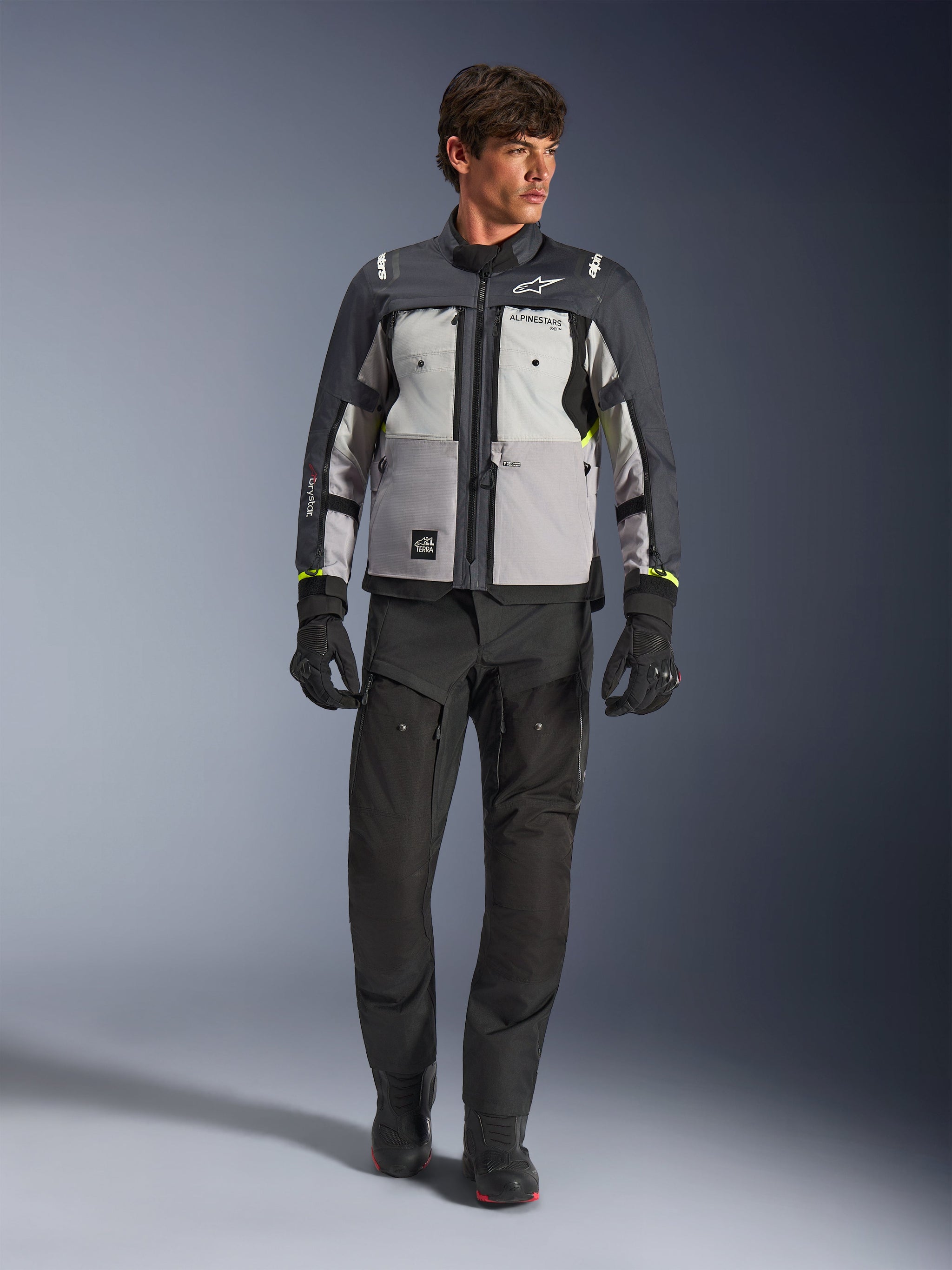 Alpinestars Cusco Drystar® Jacket in Ice Gray, Mid Gray en Deep Gray, gedragen met een zwarte adventure motorbroek en handschoenen, textiele adventure touring jas met multi-tonale grijze panelen en neongele accenten, studio-opname van een mannelijk model.
