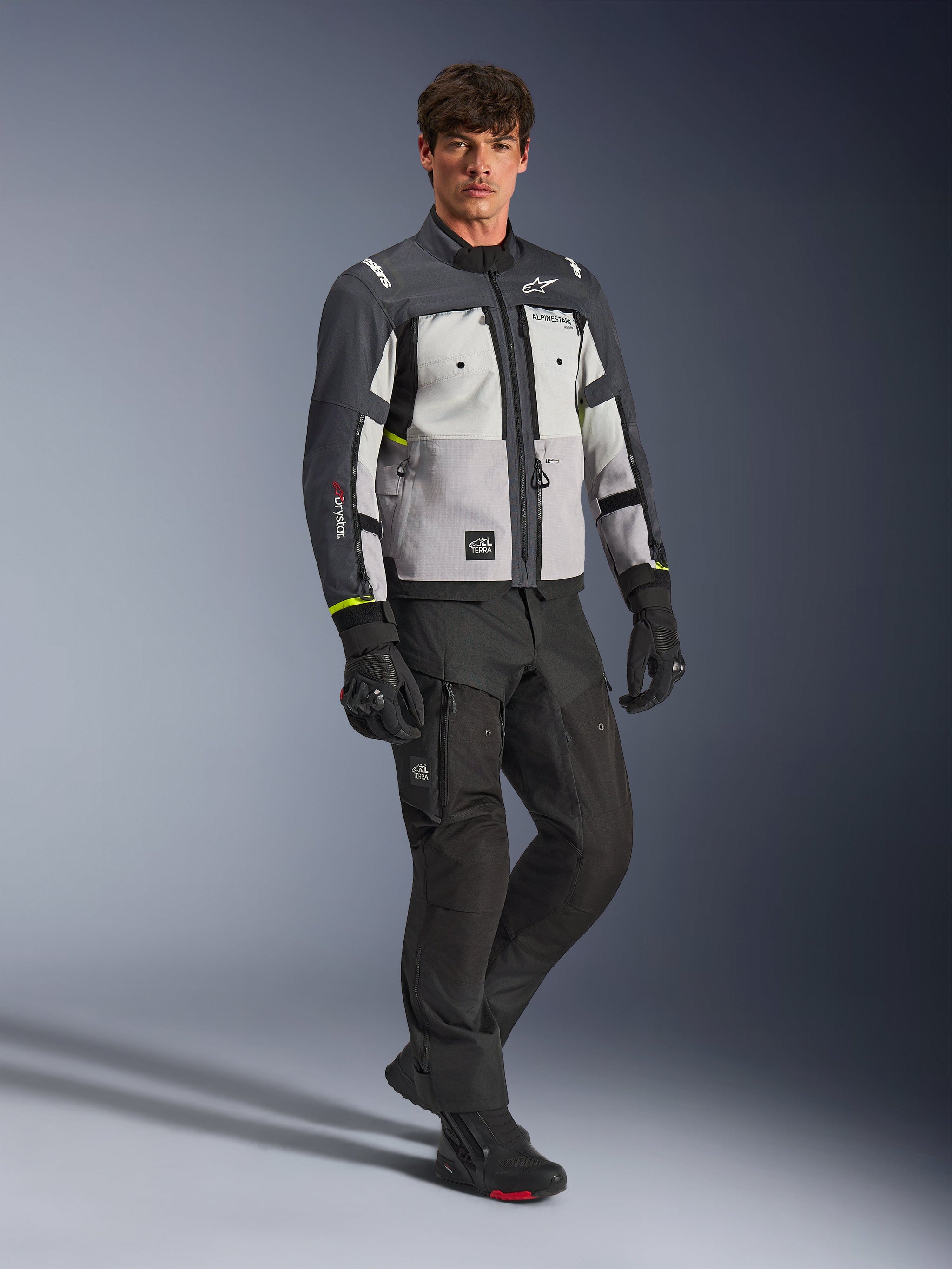 Alpinestars Cusco Drystar® jas, adventure motorjas, Ice Gray, Mid Gray en Deep Gray, gedragen door een model met zwarte broek en handschoenen, grijze panelen in verschillende tinten met een duurzame textielconstructie, voorzien van ventilatieopeningen met ritssluiting en Alpinestars-branding, professionele studiofotografie voor road- en adventure-rijuitrusting