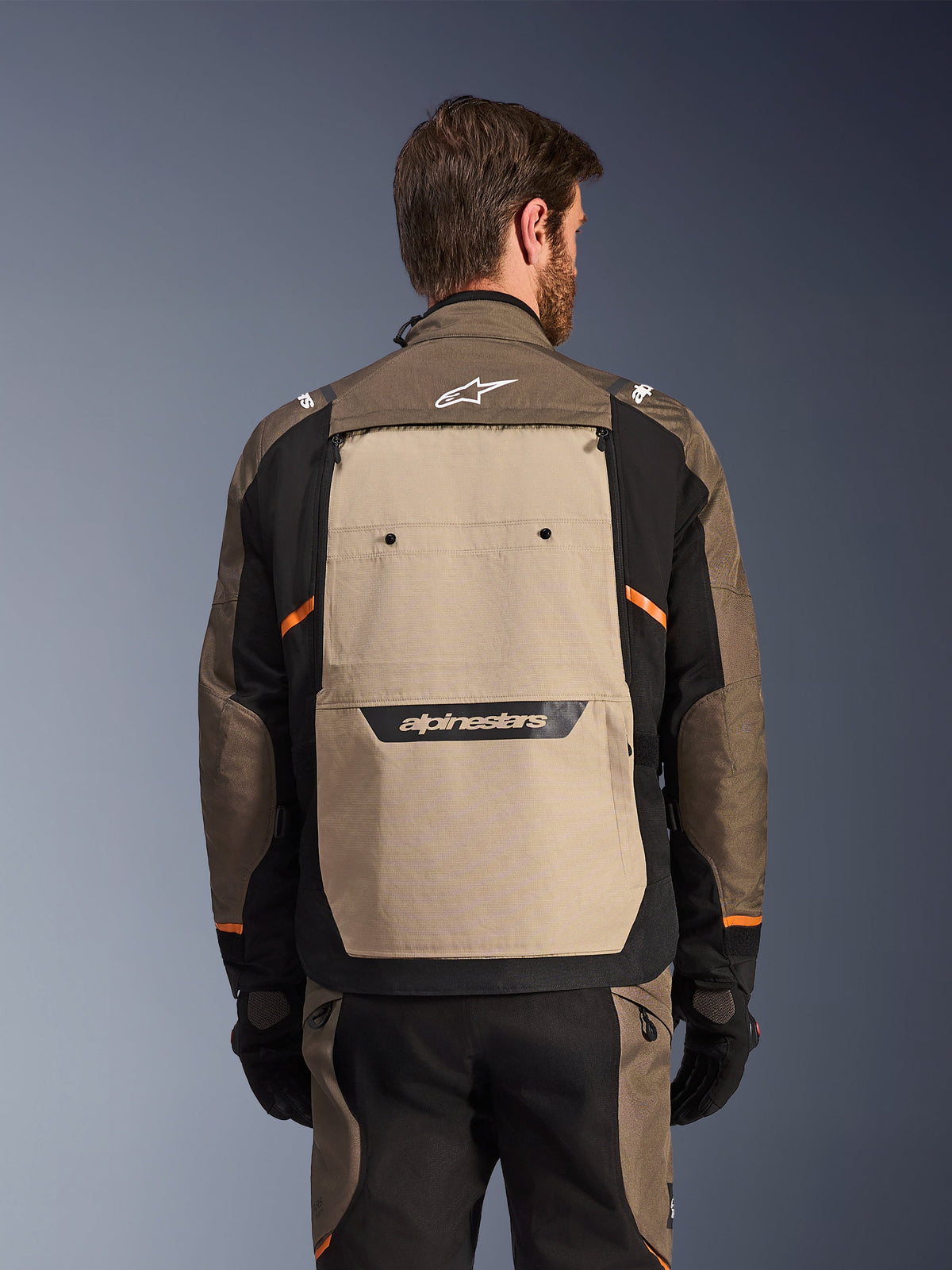 Alpinestars Cusco Drystar® Jas, Walnut Aluminum Black, achteraanzicht, voorzien van een groot beige rugpaneel met zwart logo, oranje accenten, gedragen met bijpassende broek en handschoenen, ontworpen voor adventure touring