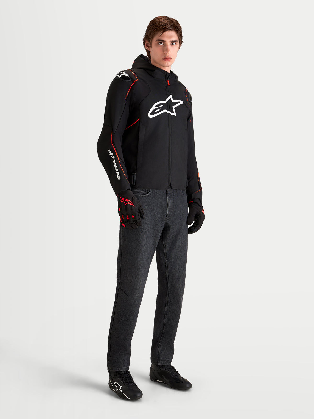 Alpinestars Zaca V2 Waterproof Jas, zwarte en rode heren motorjas met capuchon en wit borstlogo, gedragen door een mannelijk model met bijpassende motorhandschoenen en donkere jeans