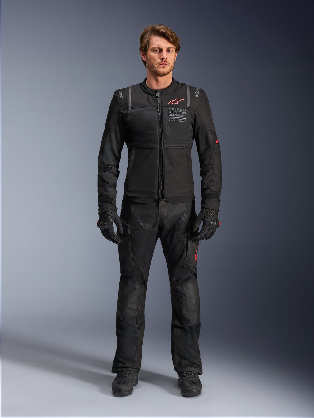 Mannelijk model met de Alpinestars ST-2 Air Jacket, zwarte motorjas met mesh-panelen en rood logo, 'Alpinestars Exploration Equipment'-tekst op de borst, gecombineerd met een zwarte technische rijbroek en handschoenen, moderne adventure touring-uitrusting