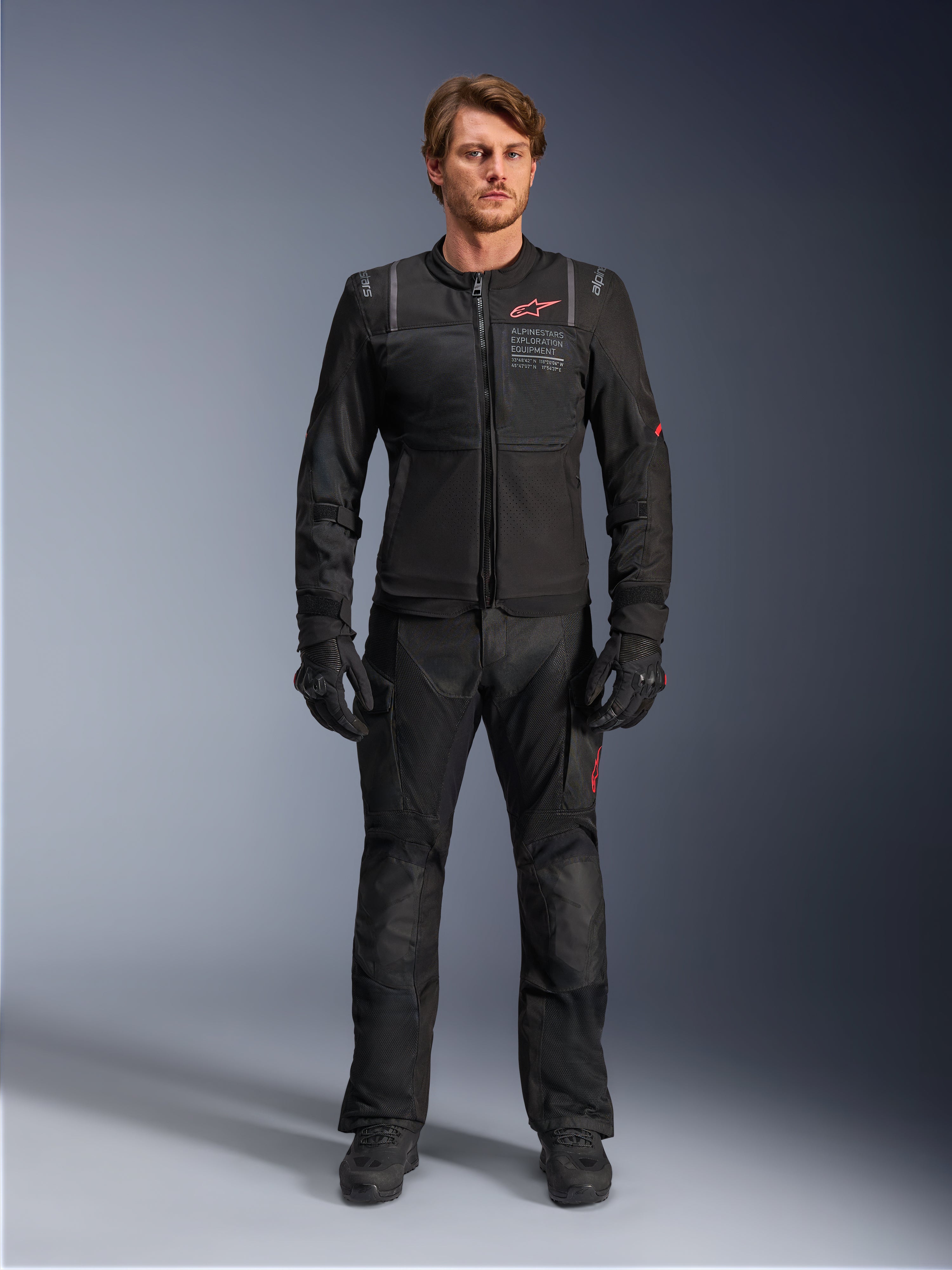 Mannelijk model met de Alpinestars ST-2 Air Jacket, zwarte motorjas met mesh-panelen en rood logo, 'Alpinestars Exploration Equipment'-tekst op de borst, gecombineerd met een zwarte technische rijbroek en handschoenen, moderne adventure touring-uitrusting