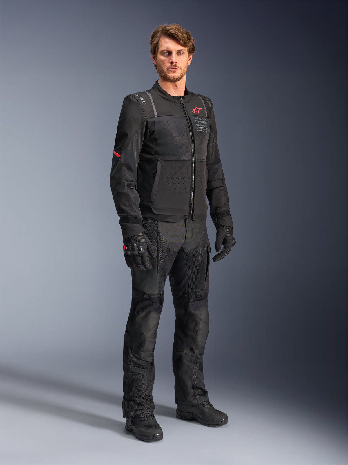 Mannelijk model in de zwarte Alpinestars ST-2 Air jas, voorzien van een rood ster-logo en 'Alpinestars Exploration Equipment' branding, gecombineerd met een bijpassende zwarte motorbroek, handschoenen en laarzen voor een moderne zomerse touring-outfit.