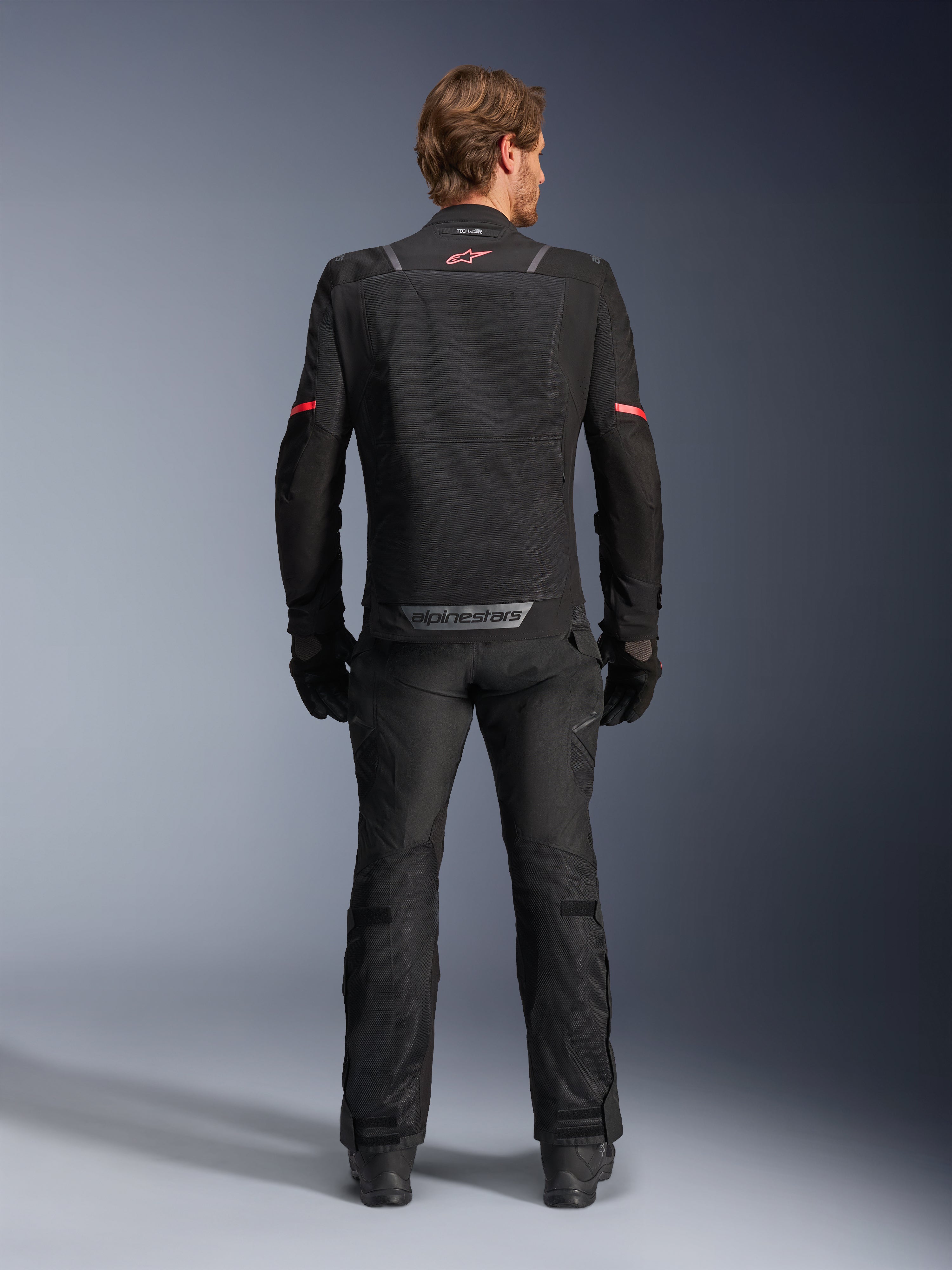 Alpinestars ST-2 Air motorjas, zwart, achteraanzicht van een motorrijder die de motorjas en bijpassende zwarte broek draagt, voorzien van mesh ventilatiepanelen, rood logo op de bovenrug en grijze Alpinestars-branding op de onderste zoom van de rug