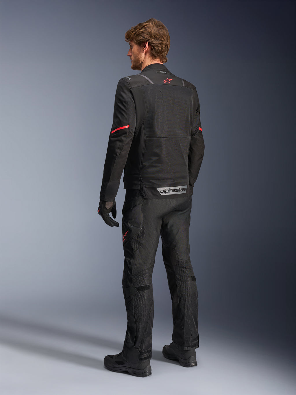 Alpinestars ST-2 Air Jacket, zwarte motorjas, achteraanzicht op model met een rood sterlogo op de bovenrug en reflecterende Alpinestars-branding bij de taille, gecombineerd met bijpassende zwarte technische motorbroek en handschoenen.