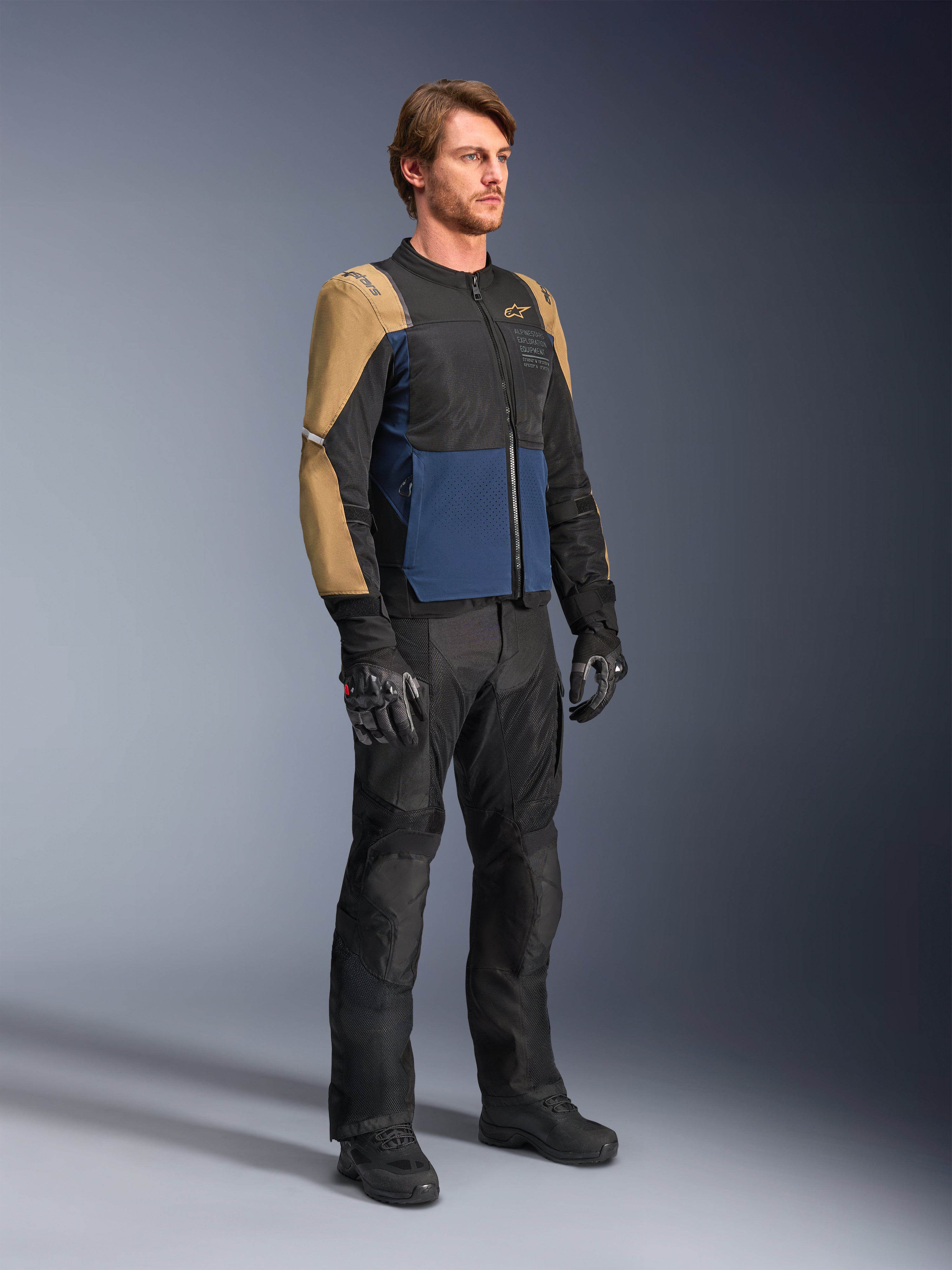 Mannelijke motorrijder die de Alpinestars ST-2 Air Jacket in Deep Blue, Dark Khaki en zwart draagt, voorzien van grote mesh-panelen en laserperforaties, gecombineerd met een zwarte motorbroek, handschoenen en laarzen voor een complete zomerse toer-look.