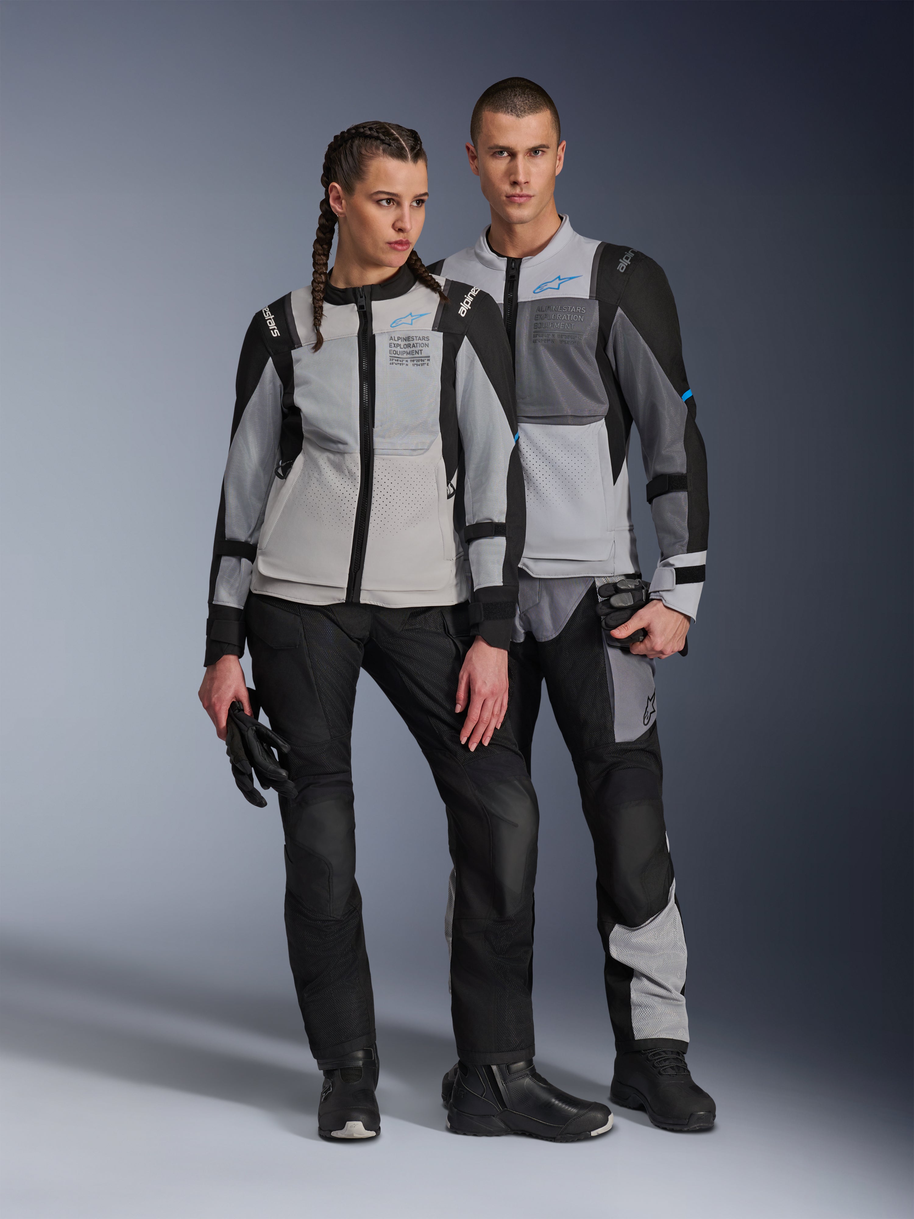 Mannelijke en vrouwelijke modellen die Alpinestars ST-2 Air-jassen dragen in Ice Gray, Dark Gray en Black, technische zomer-touringmotorkleding met grote mesh-panelen en 'exploration equipment'-branding, gecombineerd met zwarte motorbroeken, handschoenen en laarzen.