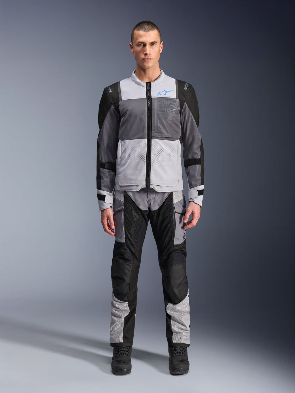 Alpinestars ST-2 Air Jas, motor-toerjas, Ice Gray, donkergrijs en zwart, gedragen door een model met bijpassende adventure-broek, voorzien van grote mesh-panelen, blauw Alpinestars-logo op de borst en Alpinestars Exploration Equipment-branding.