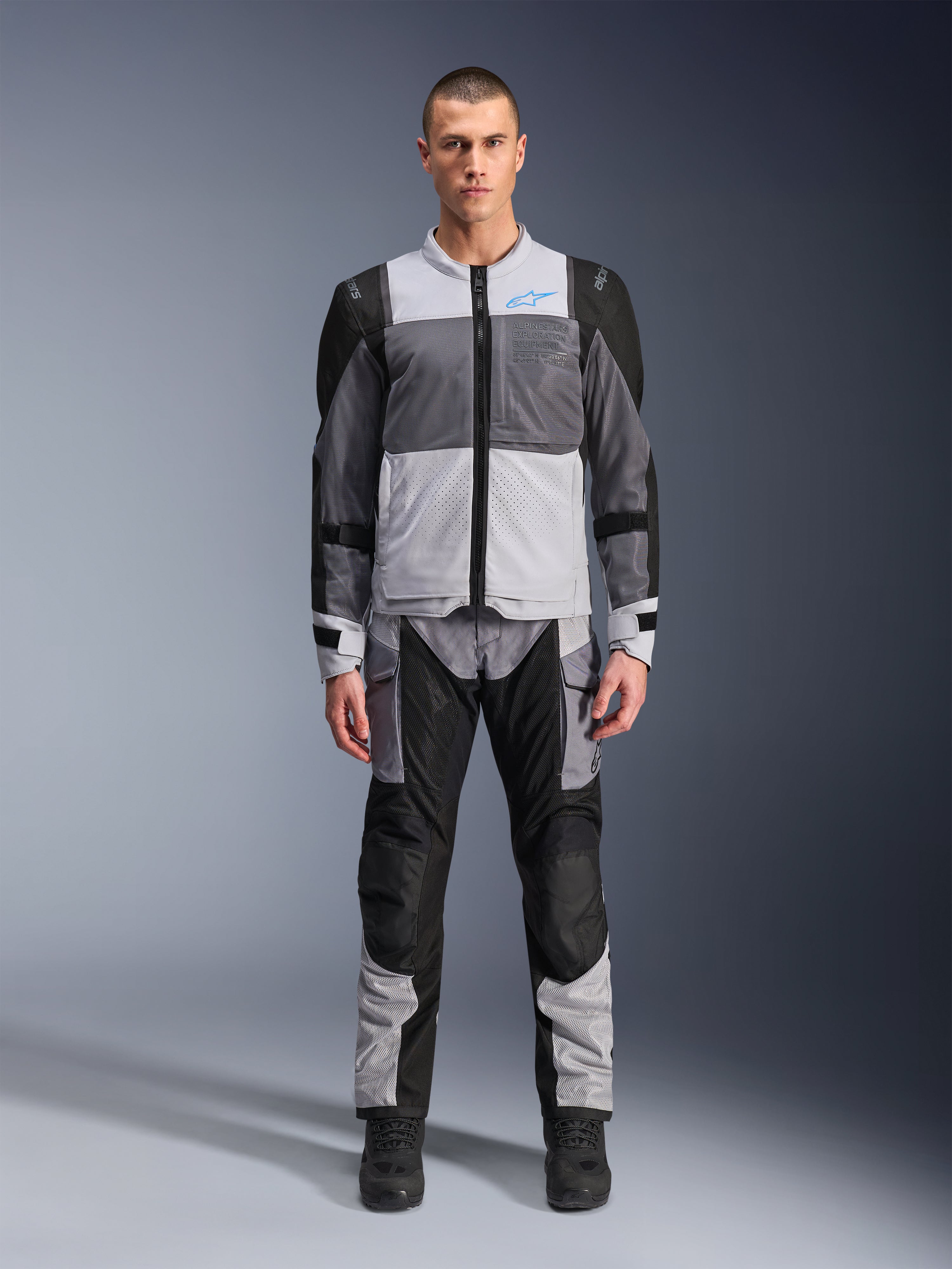 Alpinestars ST-2 Air Jas, motor-toerjas, Ice Gray, donkergrijs en zwart, gedragen door een model met bijpassende adventure-broek, voorzien van grote mesh-panelen, blauw Alpinestars-logo op de borst en Alpinestars Exploration Equipment-branding.