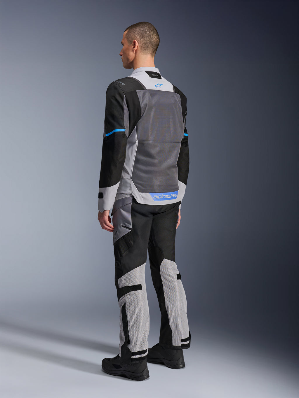 Mannelijk model van achteren die de Alpinestars ST-2 Air Jas draagt in Ice Gray, Dark Gray en Black, met een groot grijs mesh rugpaneel, blauwe logo-accenten en bijpassende motorbroek voor zomerse toertochten.