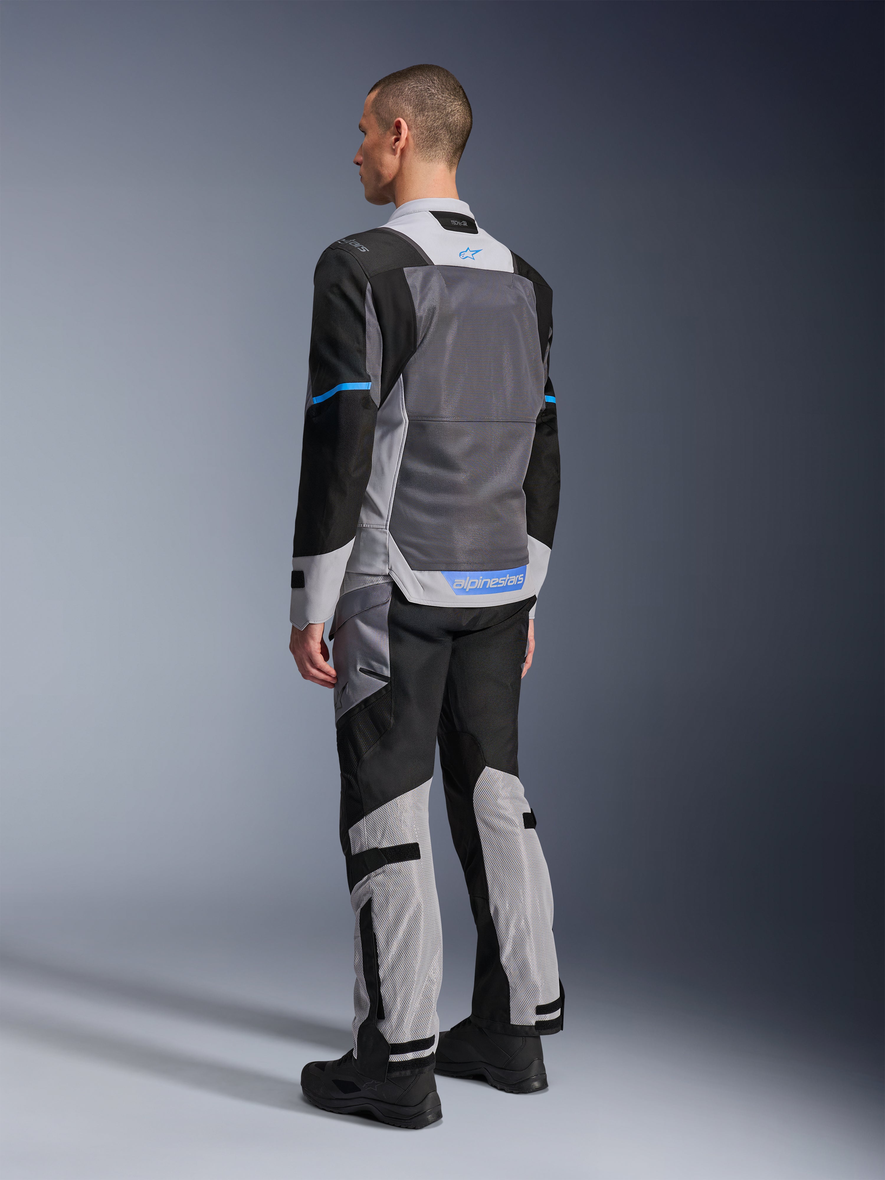 Mannelijk model van achteren die de Alpinestars ST-2 Air Jas draagt in Ice Gray, Dark Gray en Black, met een groot grijs mesh rugpaneel, blauwe logo-accenten en bijpassende motorbroek voor zomerse toertochten.