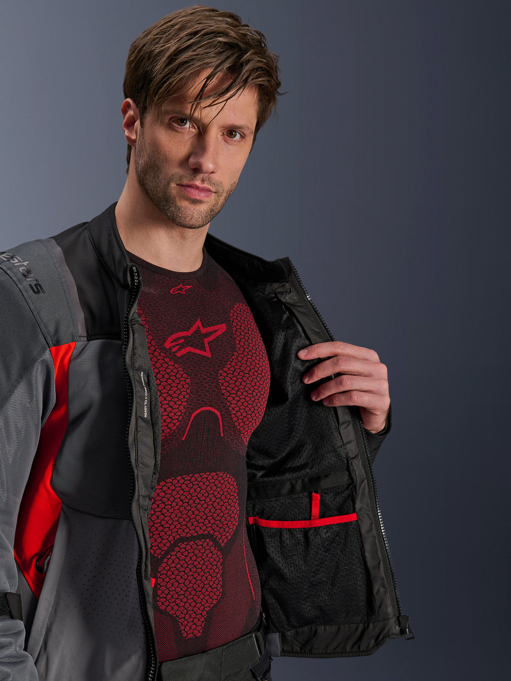 Alpinestars ST-2 Air Jas, Deep Gray Dark Gray en Bright Red, opengeritst om de interne mesh-voering en zak met rode bies te tonen, gedragen over een rode technische basislaag, moderne adventure motor touring uitrusting