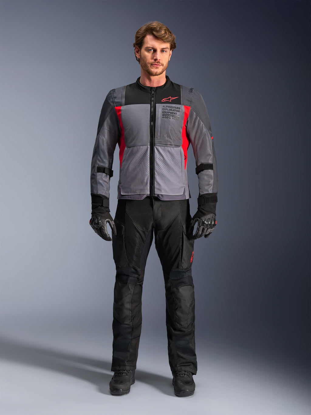 Alpinestars ST-2 Air jas in Deep Gray en Bright Red, gedragen door een mannelijk model met een zwarte motorbroek en handschoenen, voorzien van grote mesh-panelen voor luchtstroom en moderne exploration equipment branding