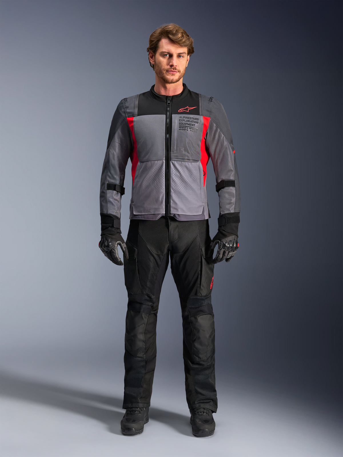 Alpinestars ST-2 Air jas in Deep Gray en Bright Red, gedragen door een mannelijk model met een zwarte motorbroek en handschoenen, voorzien van grote mesh-panelen voor luchtstroom en moderne exploration equipment branding