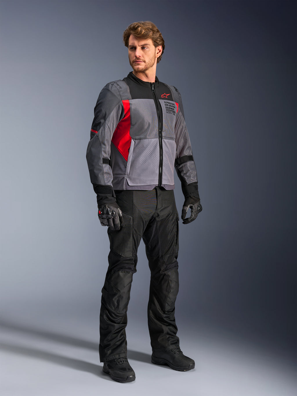 Mannelijk model draagt Alpinestars ST-2 Air Jacket, textiele motorjas, diepgrijs, donkergrijs en felrood, gecombineerd met zwarte broek, handschoenen en laarzen, voorzien van grote mesh-panelen, Alpinestars Exploration Equipment-branding en een moderne touring-esthetiek