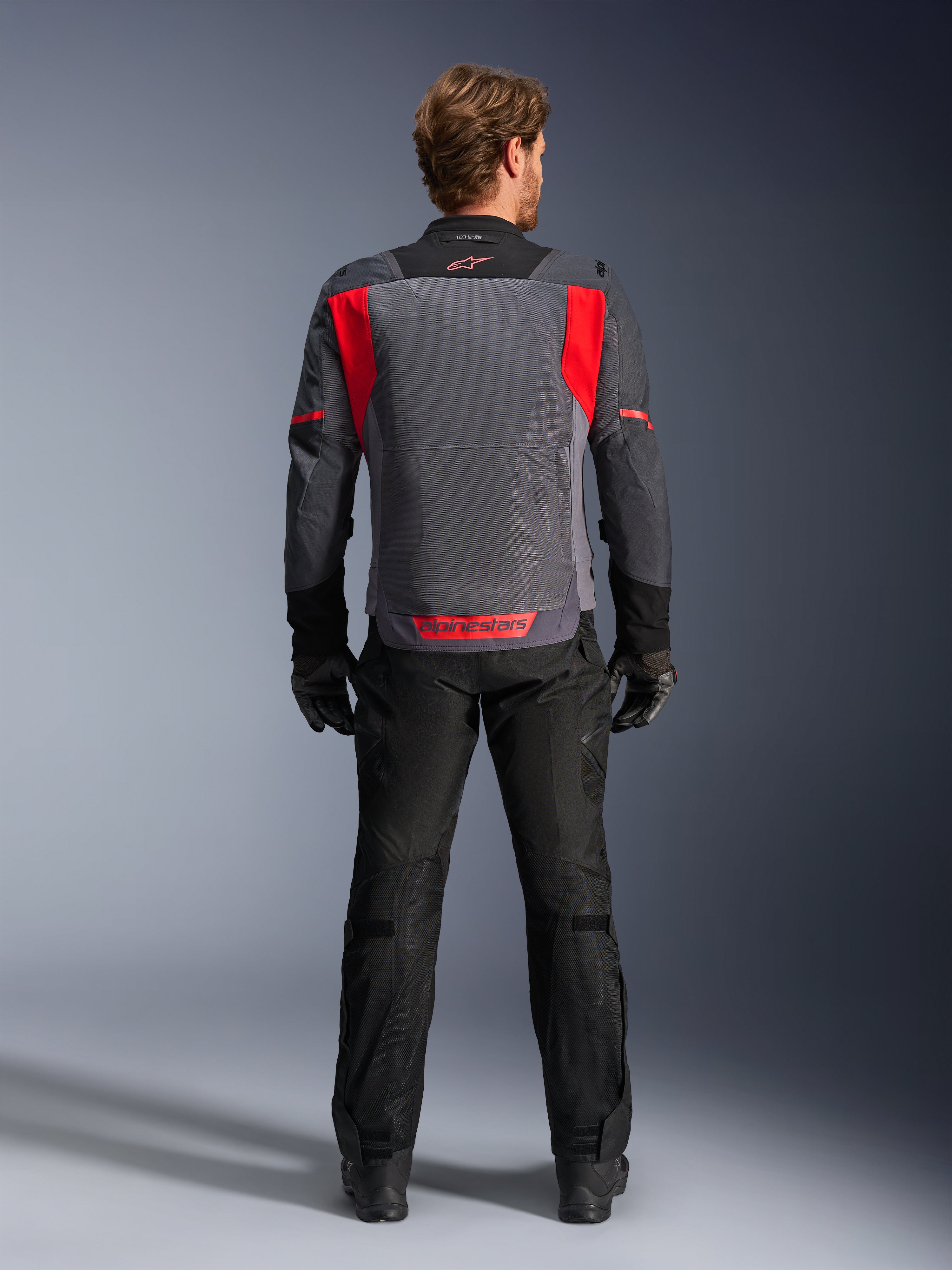 Alpinestars ST-2 Air Jas, Deep Gray Dark Gray en Bright Red, achteraanzicht gedragen door een rijder met zwarte broek en handschoenen, voorzien van grote mesh-ventilatiepanelen en rood Alpinestars-logo op de onderrug, modern zomers touring-ontwerp