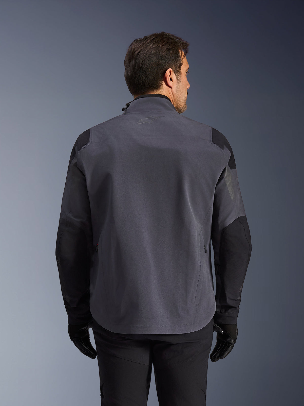 Alpinestars Maxdura Drystar®XF Laminated Armored Shell, technische adventure motorjas, donkergrijs en zwart, achteraanzicht met versterkte schouder- en elleboogpanelen, subtiel logo op de bovenrug, gedragen met zwarte handschoenen en broek, ontworpen voor bescherming in alle weersomstandigheden